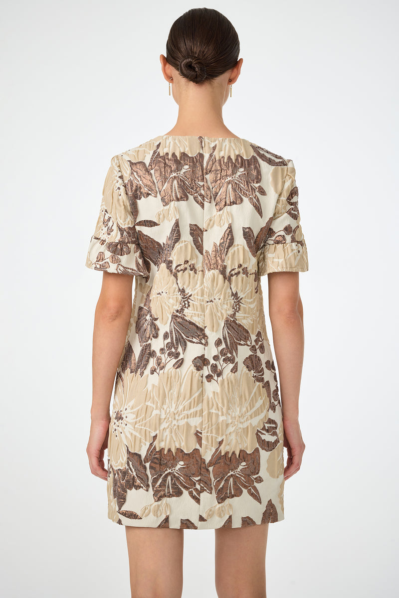 A woman with dark hair in a low bun is shown from the back against a white backdrop, wearing the Darling Flounce Sleeve Dress—an above-the-knee, short-sleeved dress with a beige and brown floral pattern on a light background.