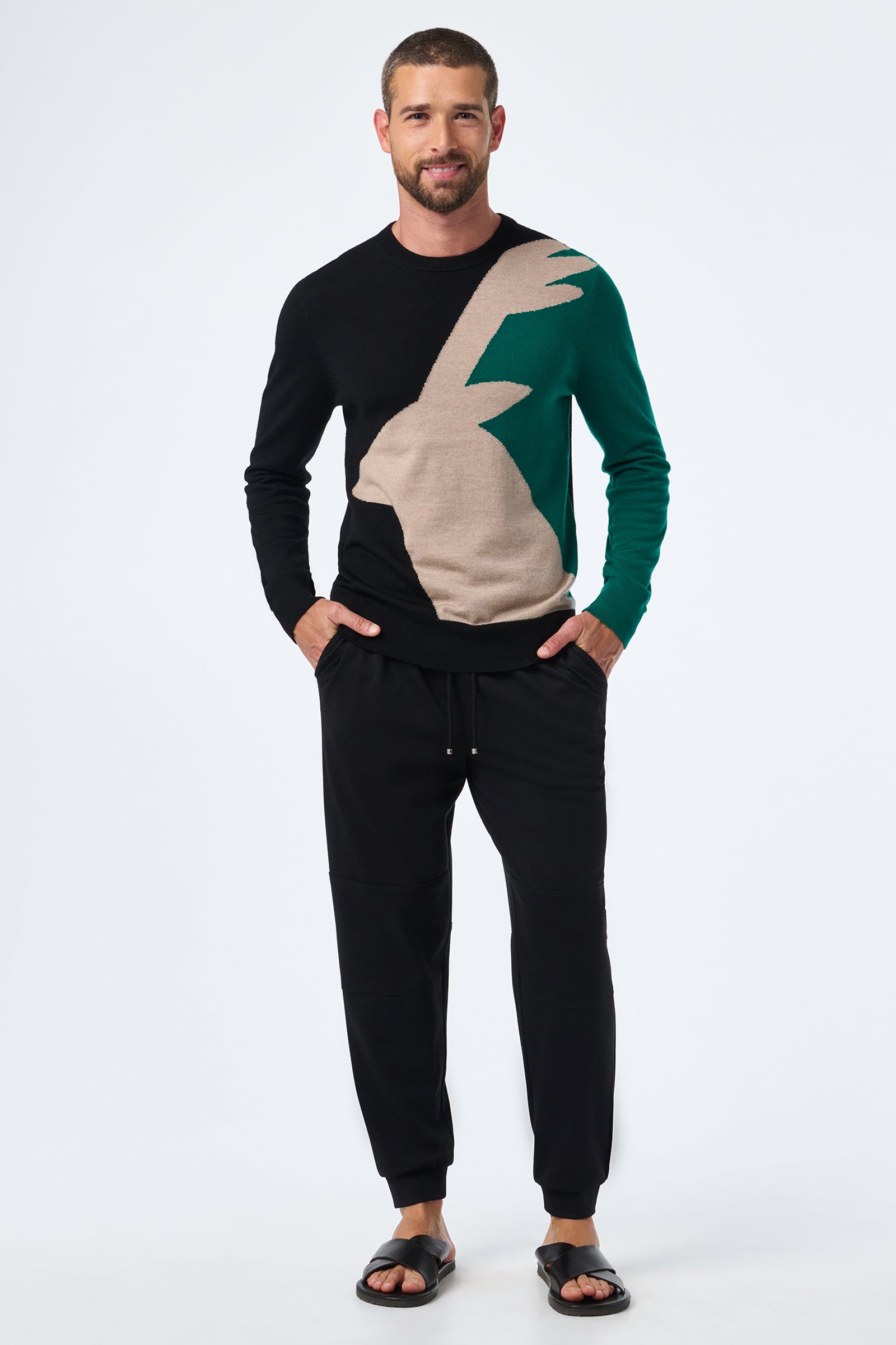A man smiles with his hands in his pockets, wearing the Hideaway Jogger, black slide sandals, and a color-block sweater featuring abstract black, green, and beige shapes. He stands against a plain white background.