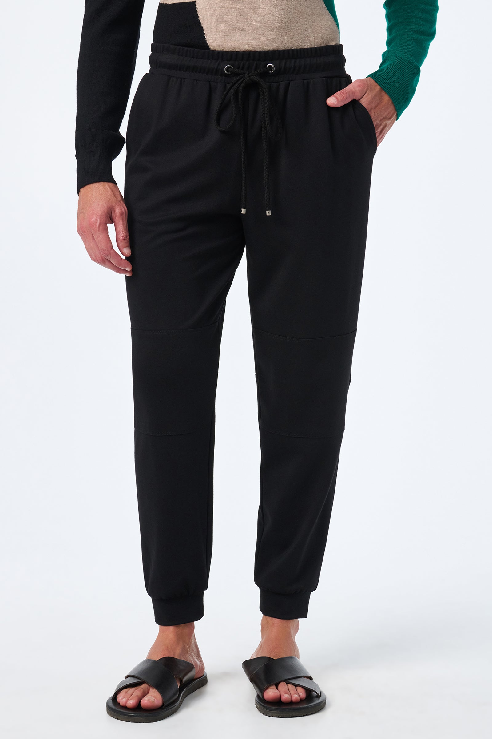 Someone wearing the Hideaway Jogger—black joggers with a drawstring waist and side pockets—paired with black slide sandals and a color-block sweater, stands against a white background.