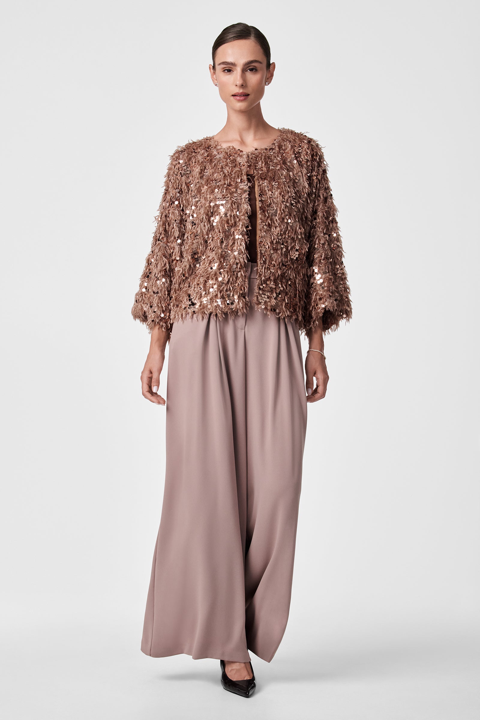 A woman stands against a plain background, wearing Saige Wide-Leg Pants in taupe, paired with a textured, sparkly rose gold jacket and black pointed heels. Her hair is neatly pulled back.