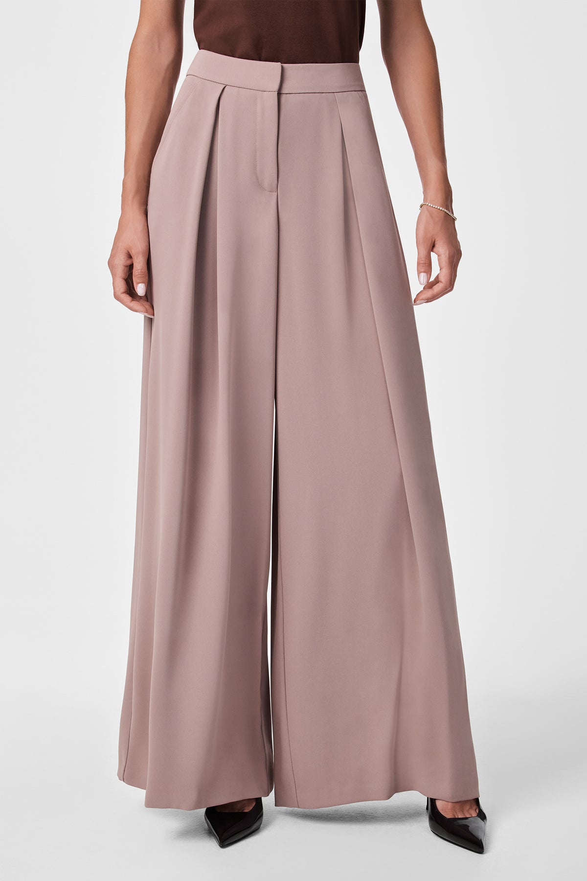A person wearing Saige Wide-Leg Short Pants in taupe, featuring high waist, pleats, front zipper, and seam, paired with a dark top and black pointed-toe shoes. Only their lower torso and legs are visible.