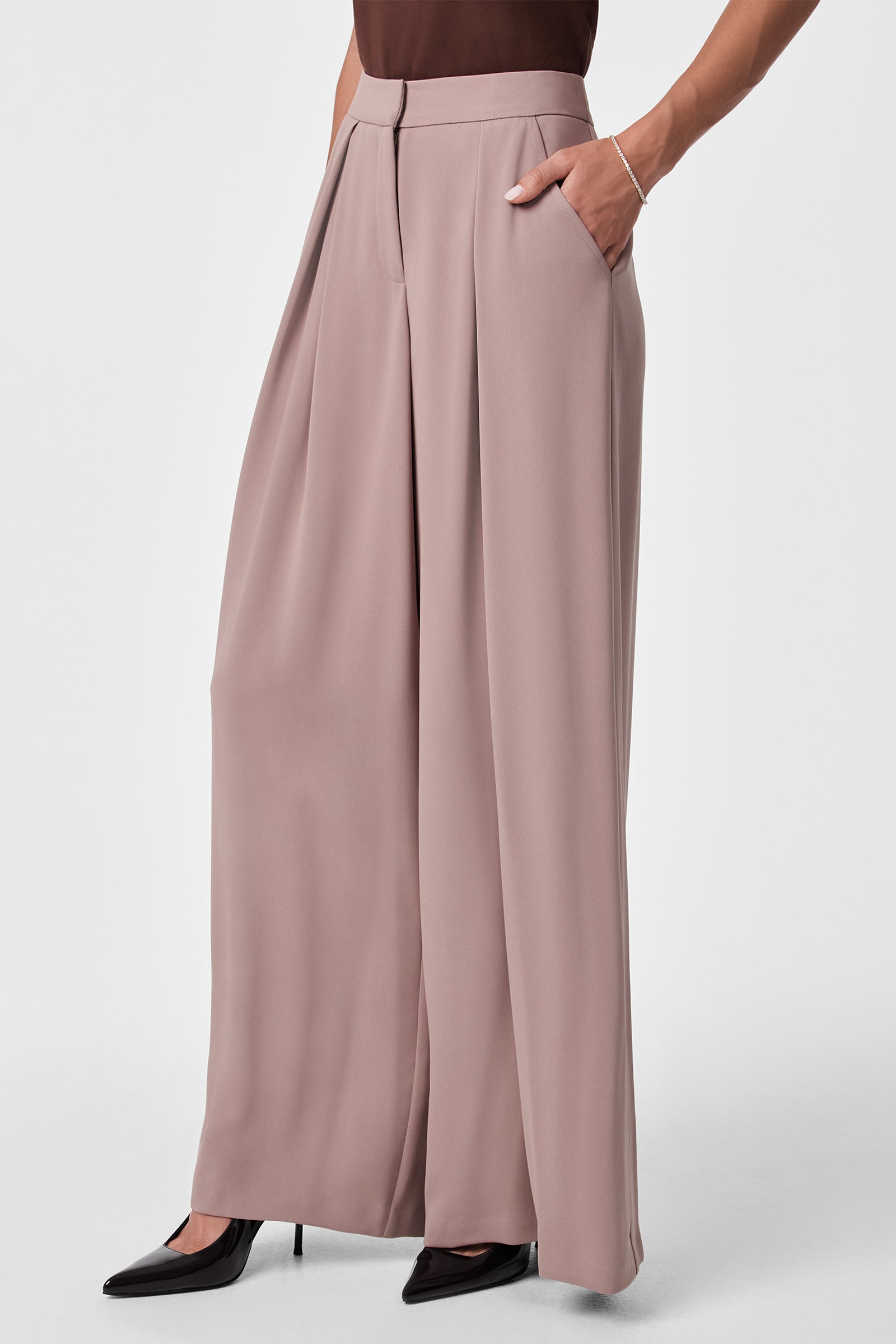 A person wears Saige Wide-Leg Pants—high-waisted, pleated taupe trousers—with a brown top and black pointed-toe heels. One hand is in a pocket; only the lower body appears against a plain background.