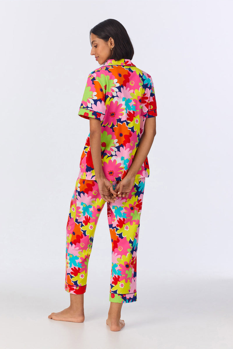 A person stands barefoot facing away, wearing the Crop Classic PJ Set in Pop Floral Stretch Jersey. The bright pajamas feature a bold pink, red, yellow, green, and white floral pattern. Their hands are clasped behind their back against a white background.