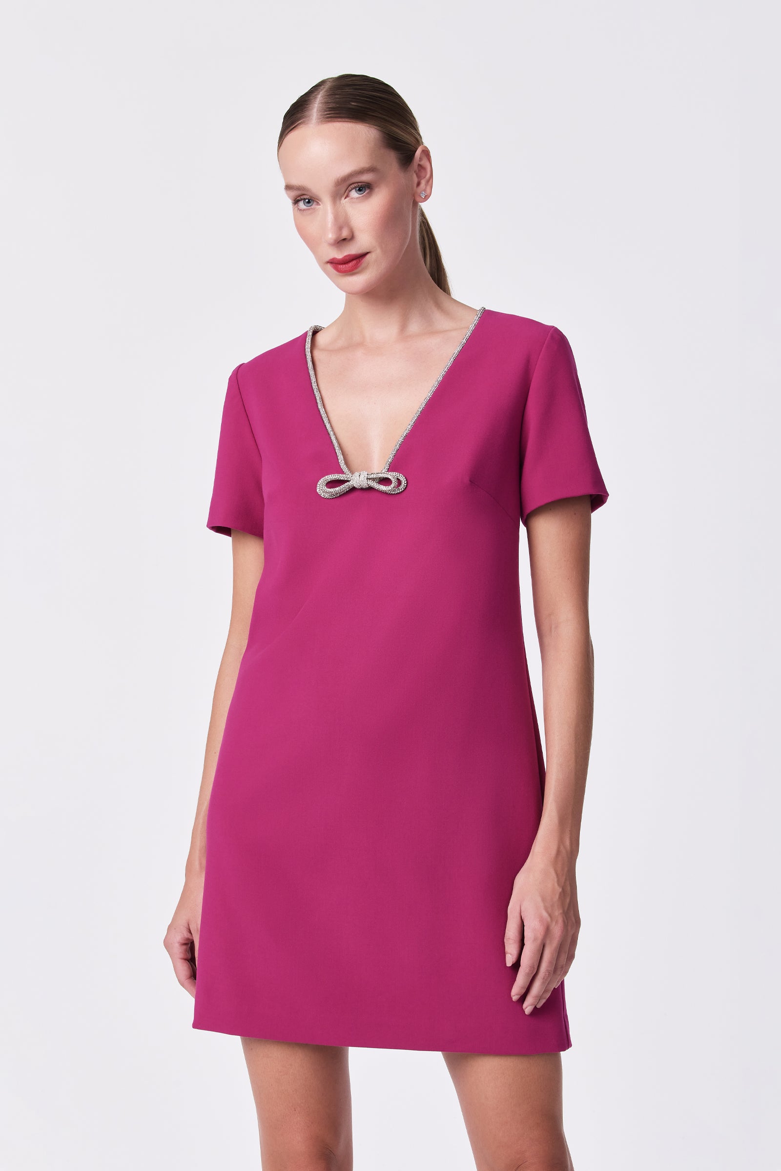A woman wears the Obelle Dress—a short-sleeved, magenta style with a deep V-neckline and decorative center bow—standing against a white background and smiling subtly at the camera.