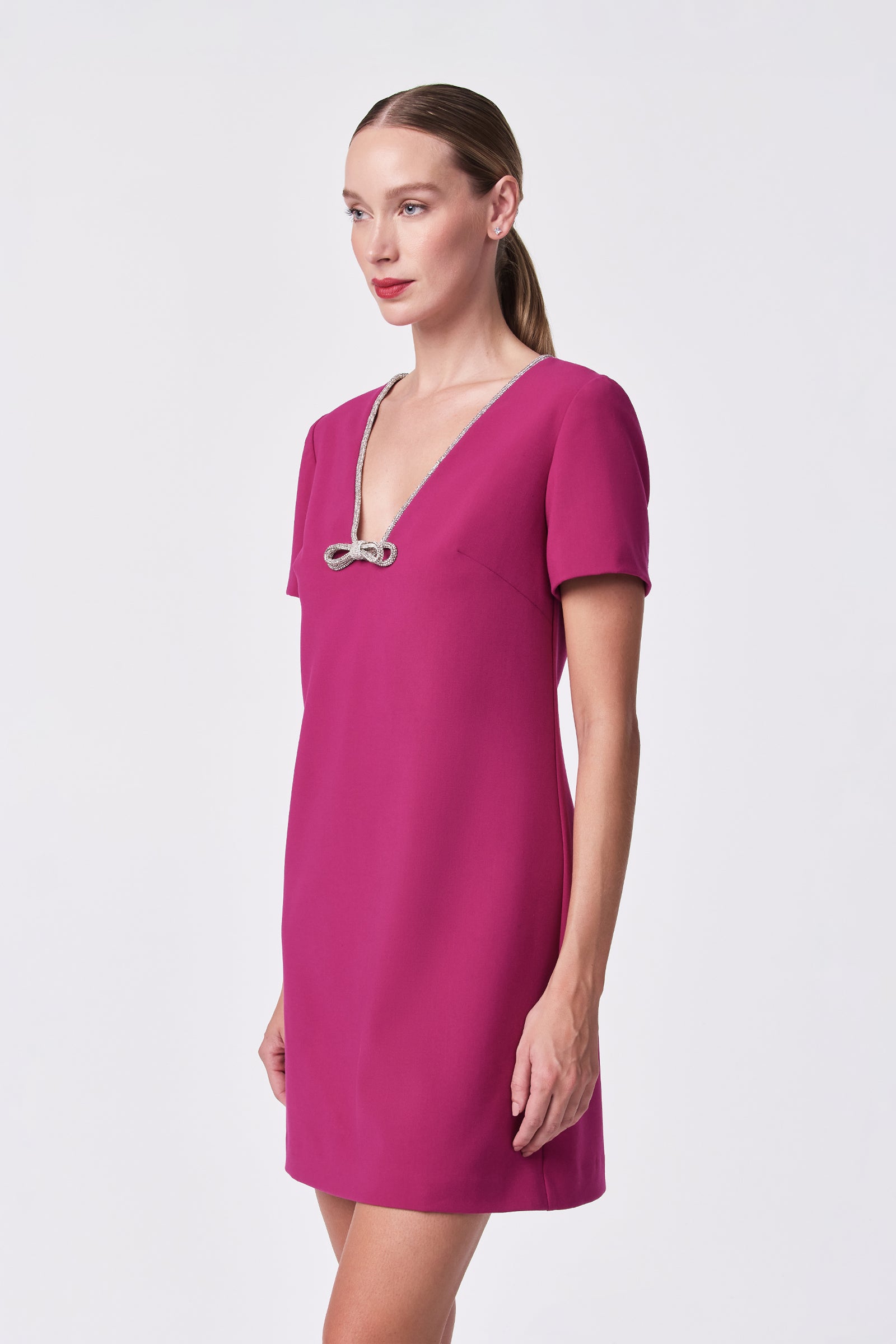 A woman models the Obelle Dress—a short-sleeved, magenta design featuring a deep V-neckline and a decorative metallic bow—standing against a plain white background and gazing slightly to the side.