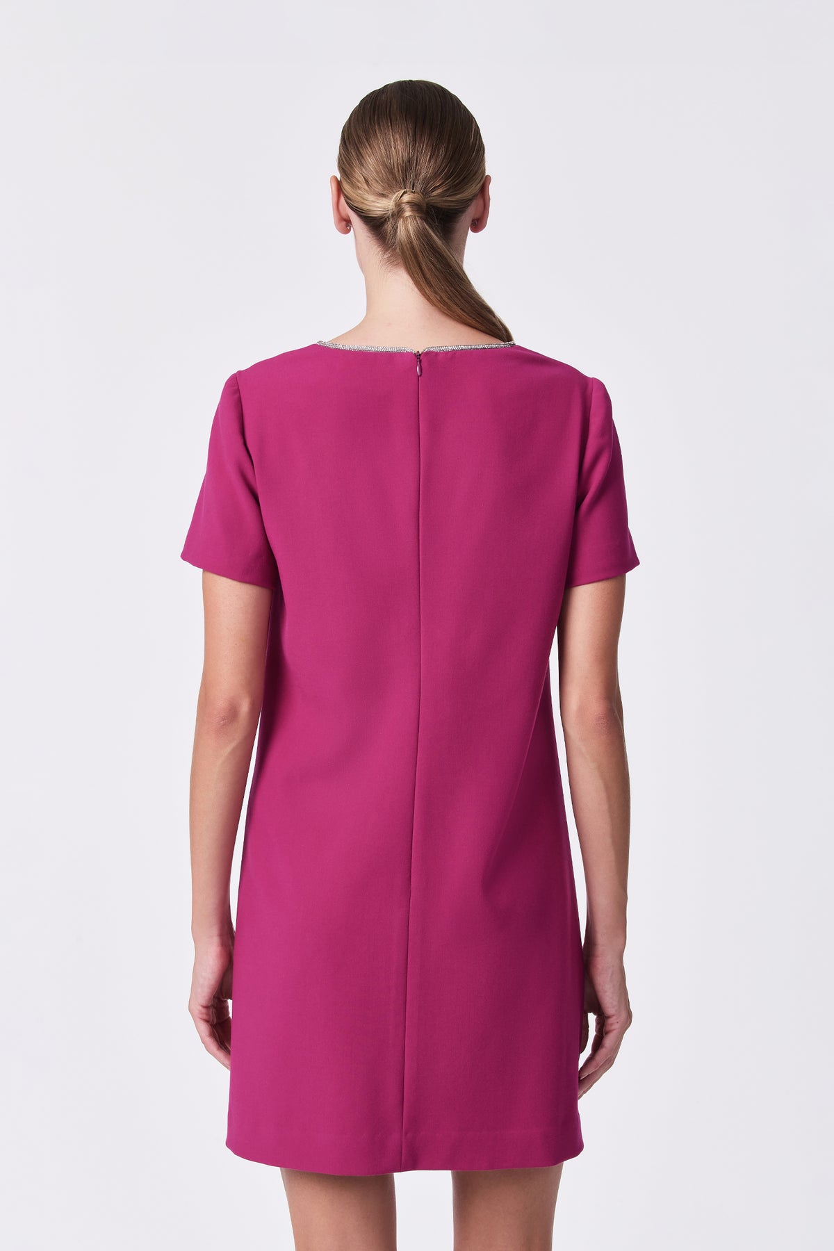 A woman with straight, light brown hair in a low twisted ponytail is shown from the back, wearing the short-sleeved, knee-length magenta Obelle Dress, standing against a plain white background.