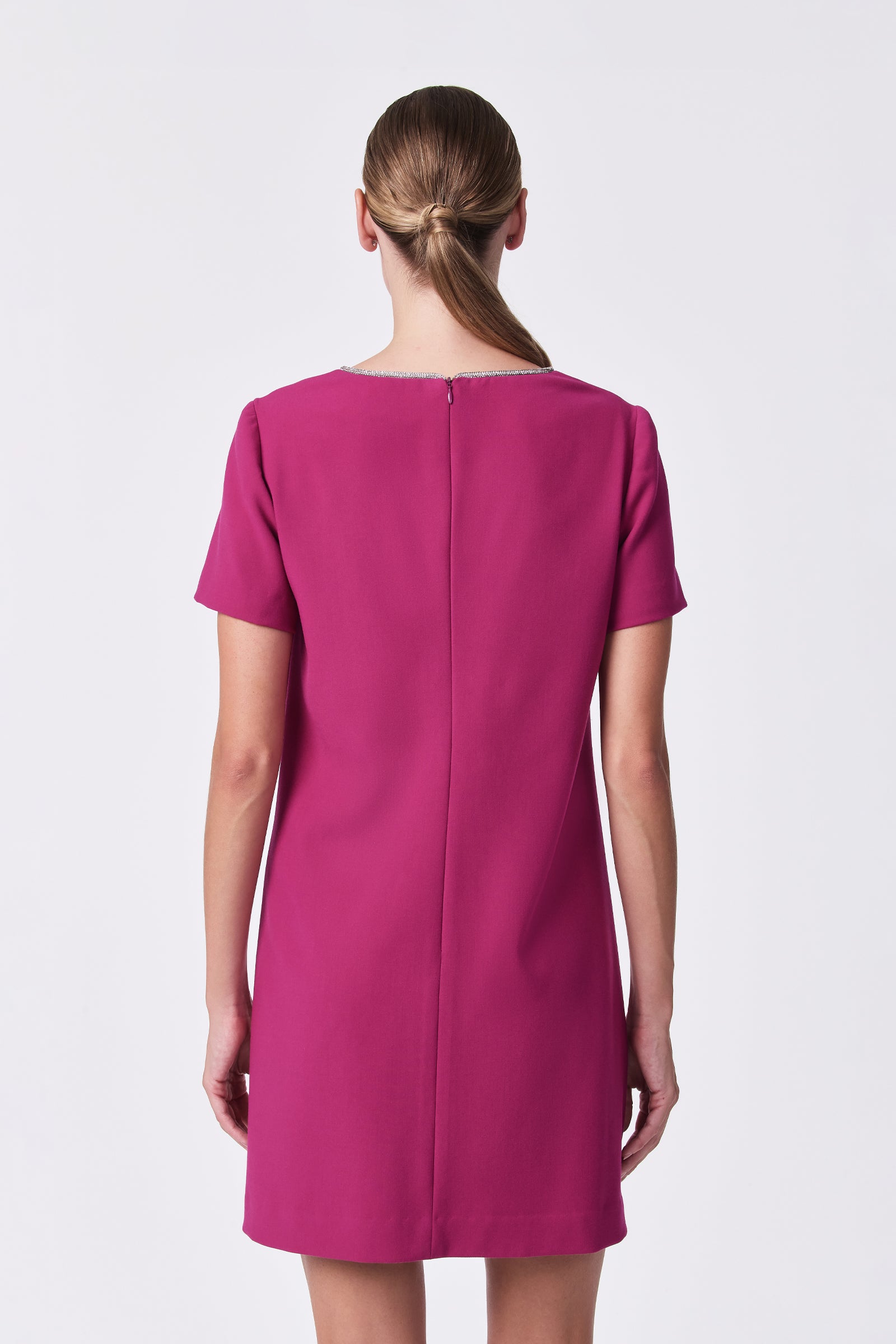 A woman with straight, light brown hair in a low twisted ponytail is shown from the back, wearing the short-sleeved, knee-length magenta Obelle Dress, standing against a plain white background.