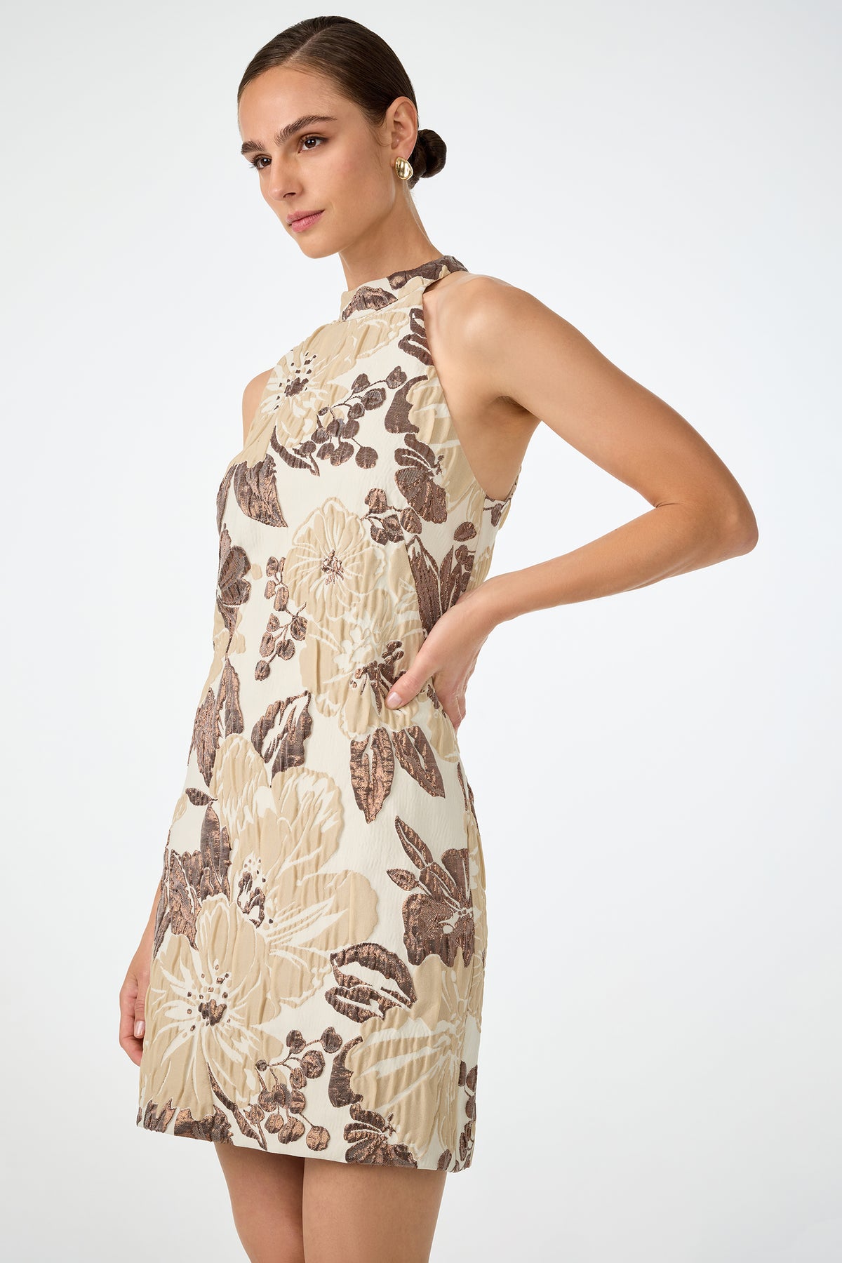 A woman wears the Garam Sleeveless Halter Dress, a cream mini dress with large beige and brown floral patterns, standing against a plain white background.