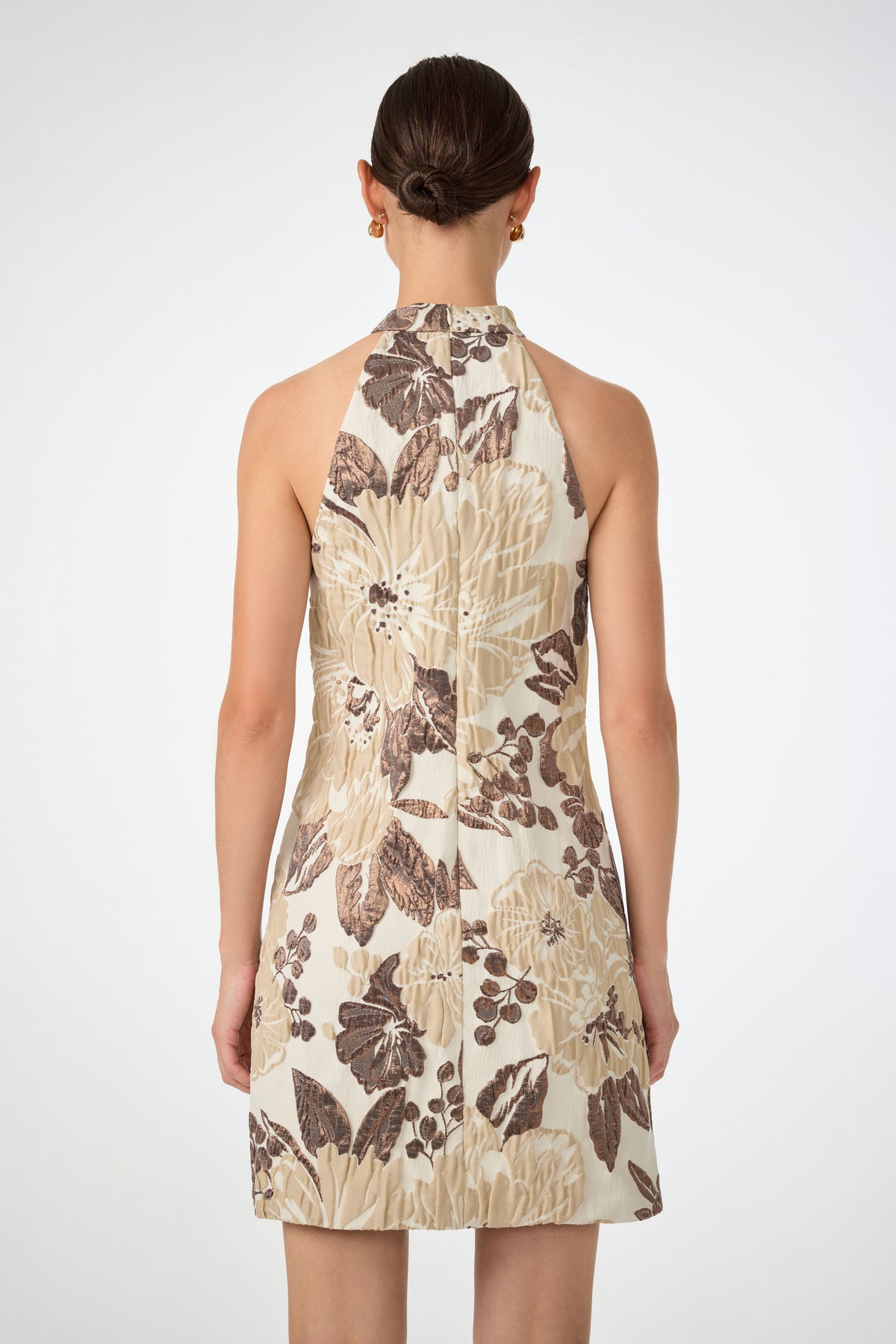 A woman with dark hair in a low bun models the Garam Sleeveless Halter Dress, featuring a beige and brown floral pattern, shown from the back against a plain white background.