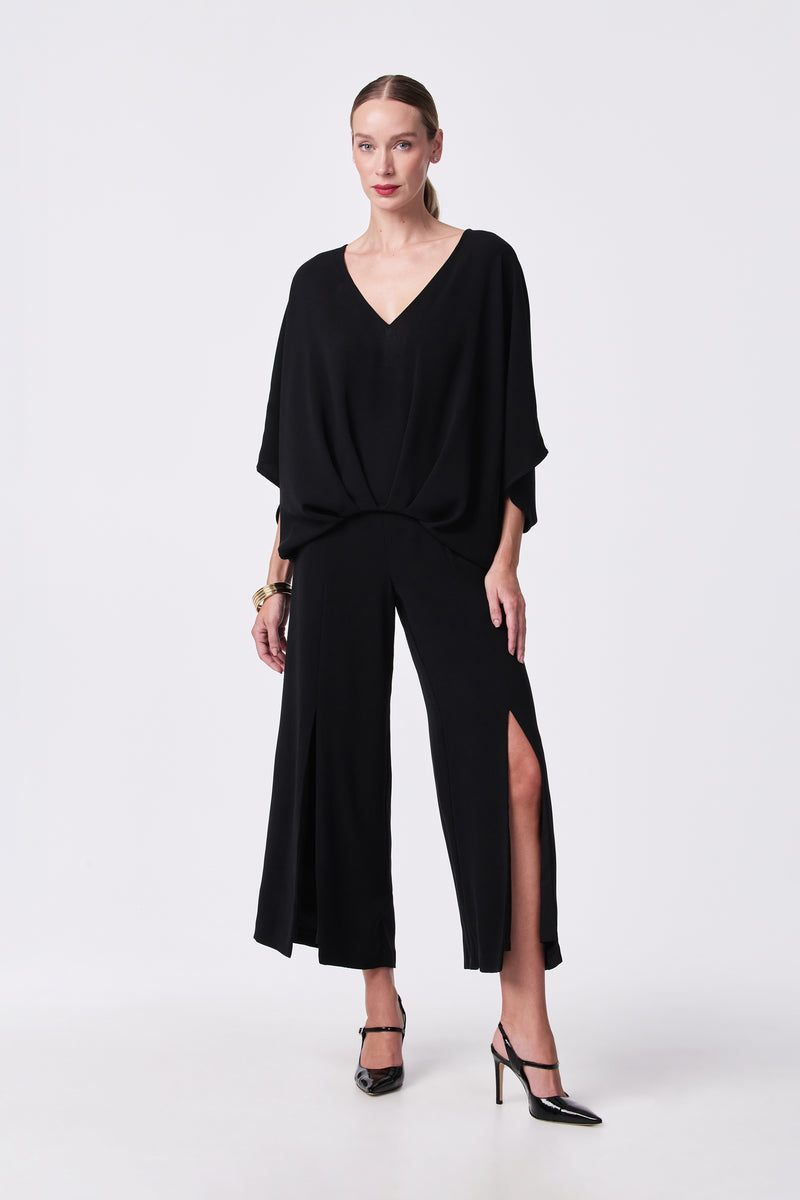 A woman stands against a white background wearing the Davon Pant, a wide-leg black pant with thigh-high slits, paired with a loose black V-neck top. She has her hair pulled back and accessorizes with black heels and gold jewelry.
