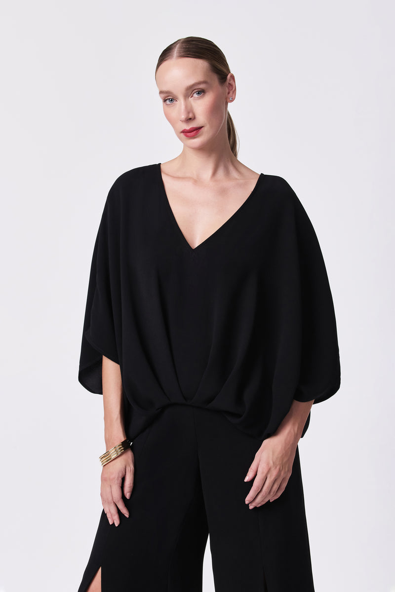 A woman with straight, light brown hair tied back wears the Coralline 2 Top, a loose-fitting black V-neck style, along with matching black pants. She stands against a plain white background, looking at the camera with a relaxed expression.
