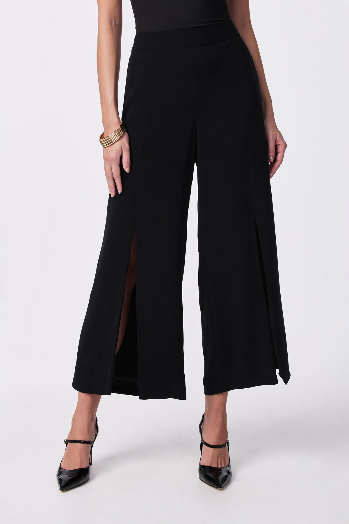 A person models the Davon Pant—wide-leg black pants with front slits—paired with a black sleeveless top, black heels, and gold bangles, standing against a plain light background.