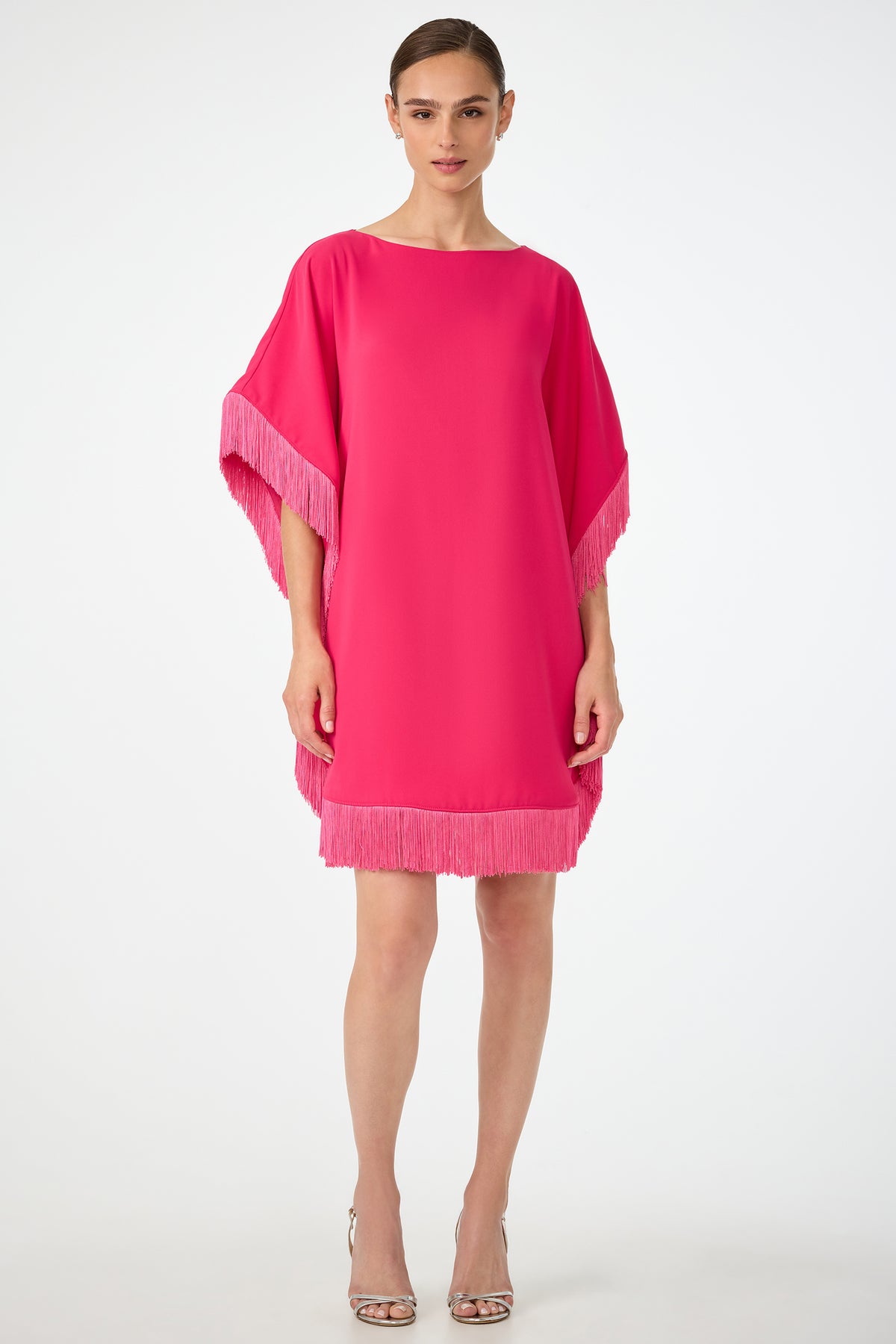 A woman wears the Mini Neena Fringe Caftan Dress, a loose-fitting bright pink dress with fringe on the sleeves and hem, paired with open-toe heels. Her hair is pulled back as she stands against a plain white background.