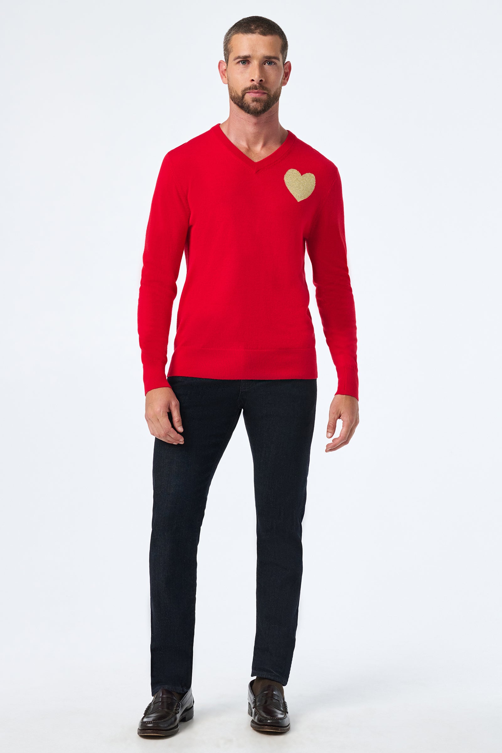 A man stands against a white background wearing the Lover Crewneck, a red sweater with a gold heart on the left chest, paired with dark pants and brown shoes.