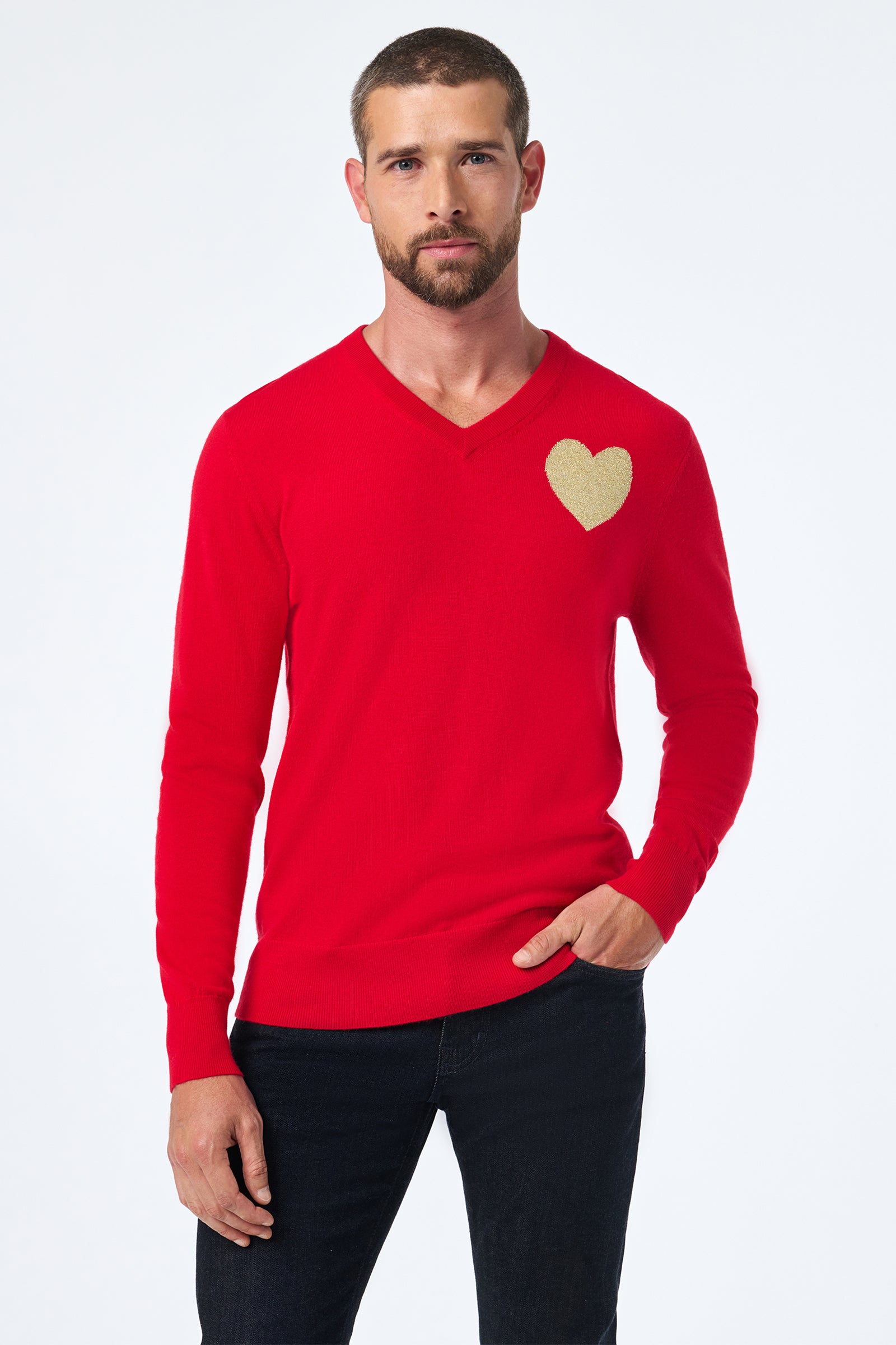 A man with short hair and a beard wears the Lover Crewneck, a bright red sweater with a beige heart on the left chest, paired with dark pants, standing against a plain white background.