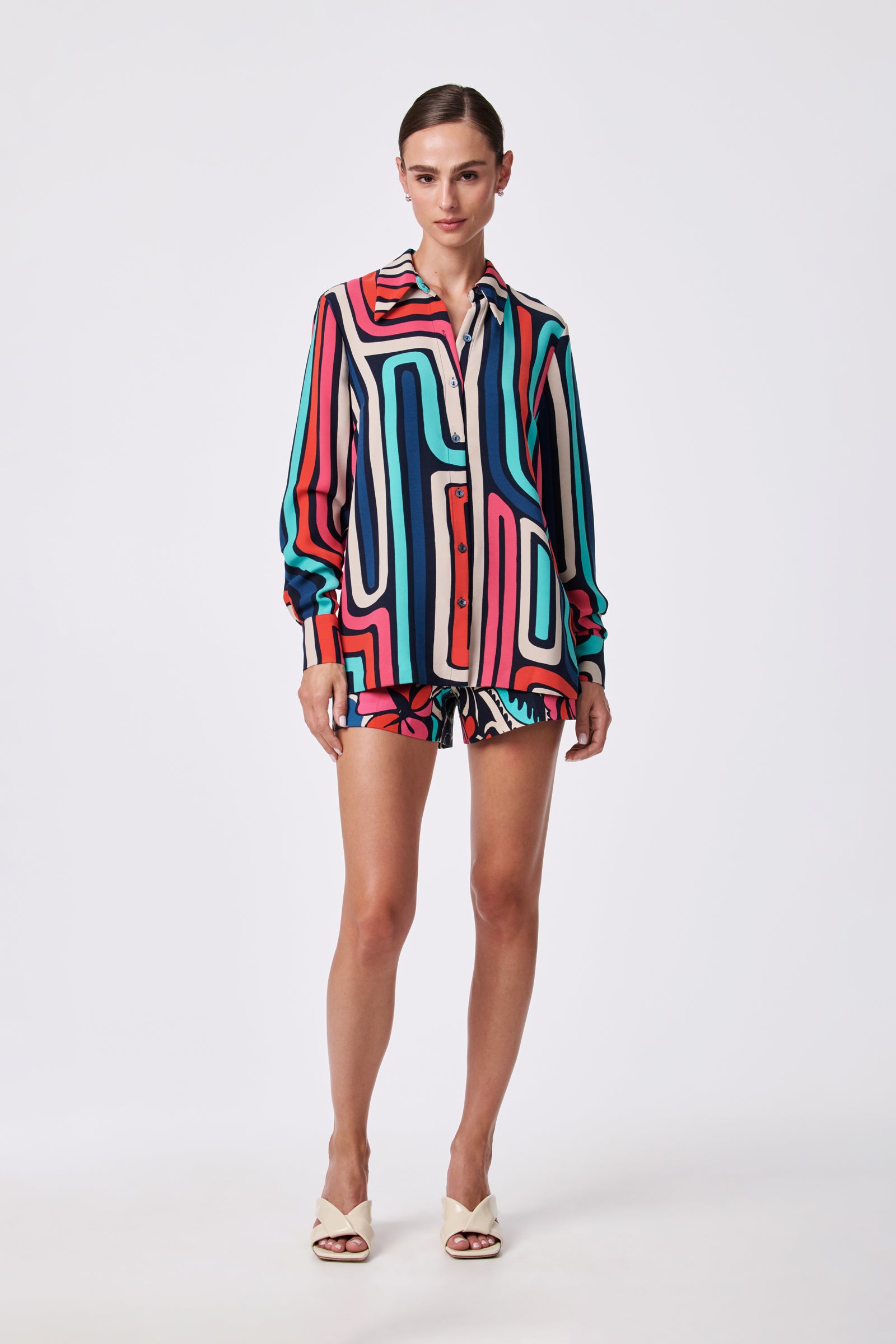 A woman wears the Montero Top, a button-up shirt with a bold geometric pattern in blue, red, black, and white. She pairs it with matching shorts and white heels, posing against a plain white background.