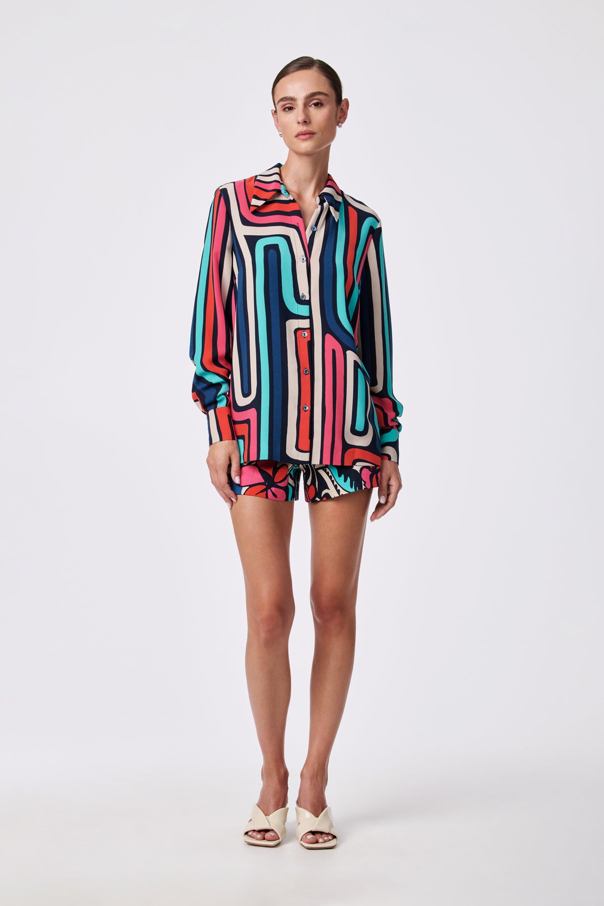 A woman poses against a white background wearing the Corbin 2 Short and a matching long-sleeve shirt, both featuring a bold geometric pattern in red, teal, black, white, and pink. She completes the look with white heeled sandals.