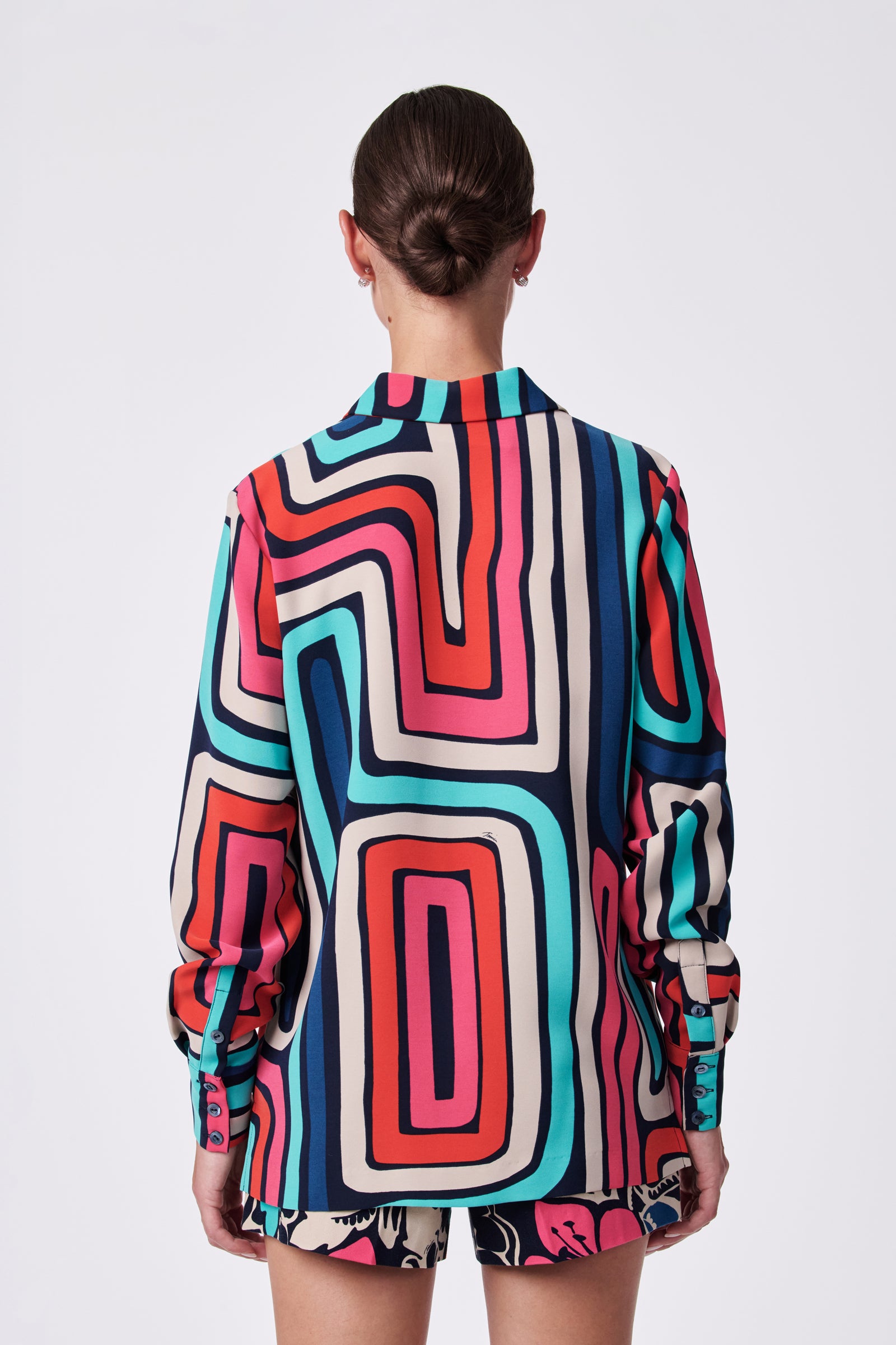 A person with hair in a low bun is seen from behind, wearing the Montero Top—a long-sleeved shirt featuring a bold geometric pattern in red, teal, navy, and cream—paired with patterned shorts against a plain white background.