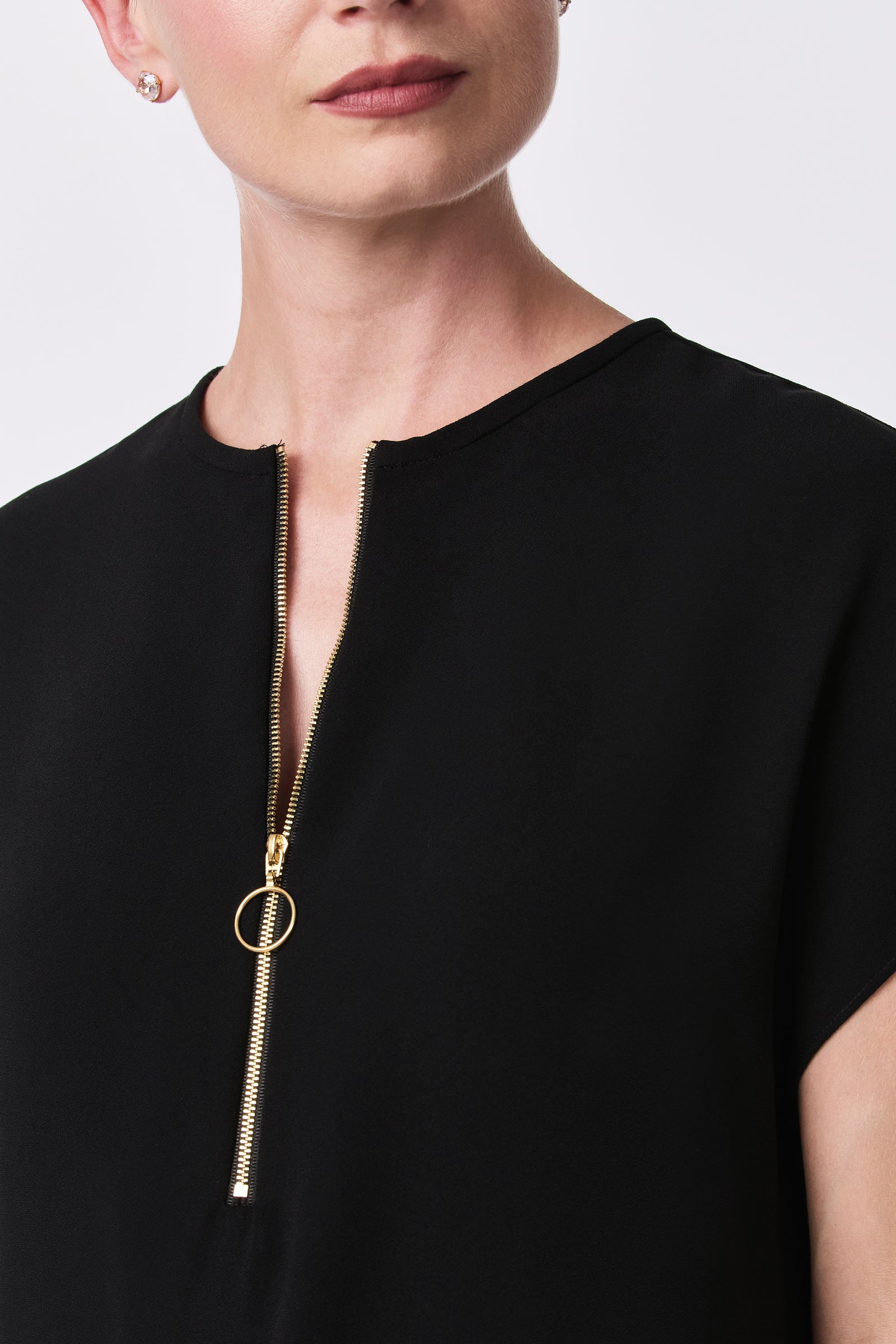 A person wears the Hasil Dress—a black top with a gold zipper and round pull ring, partially unzipped at the neckline. Only the individual's lower face and upper torso are shown; they have short hair and stud earrings.