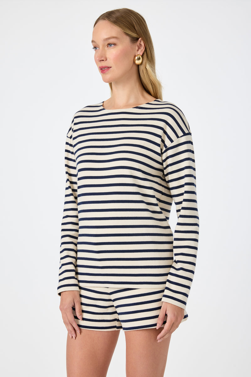 A woman wears the Novena Top in Noh Nightfall Stripe‚Äîlong-sleeve, black and white stripes‚Äîpaired with matching shorts, standing against a plain white background and gazing slightly to the side.