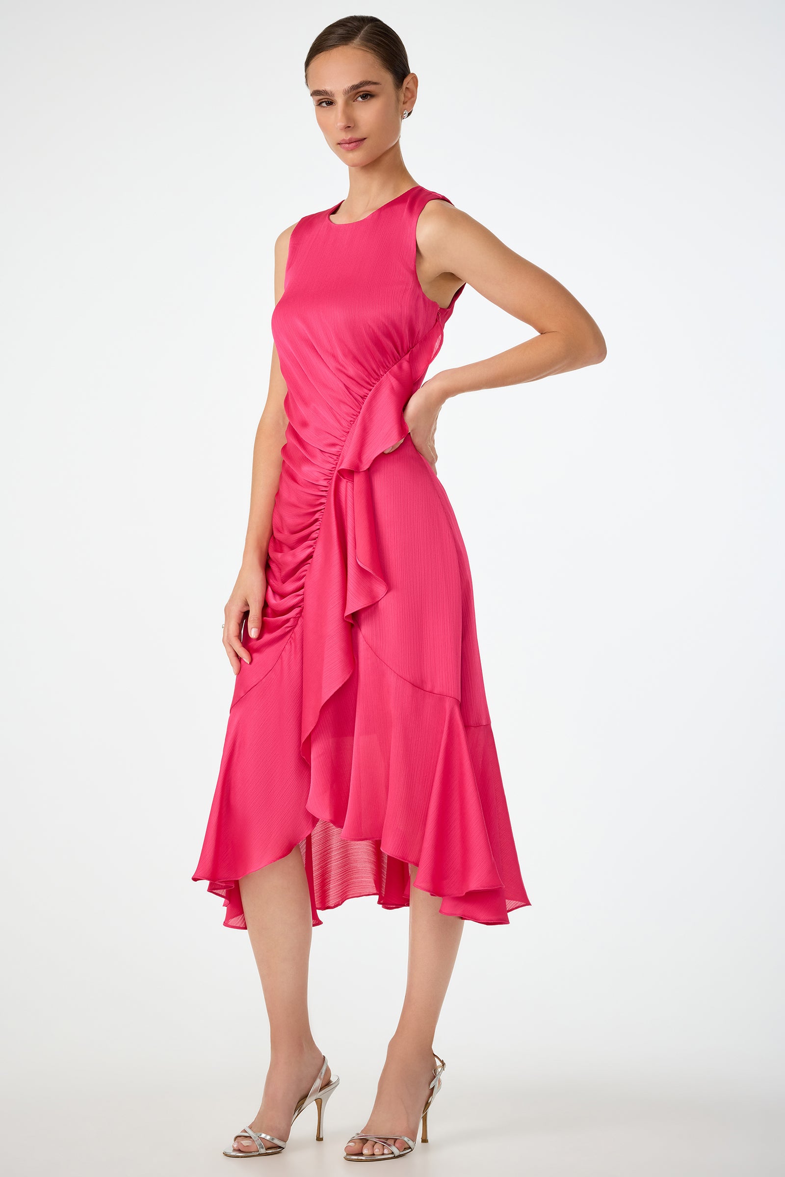 A woman poses in the Amara 2 Sleeveless Ruffle Dress, a bright pink asymmetric midi dress with ruched and ruffled details, standing with one hand on her hip and pairing it with silver high-heeled sandals.