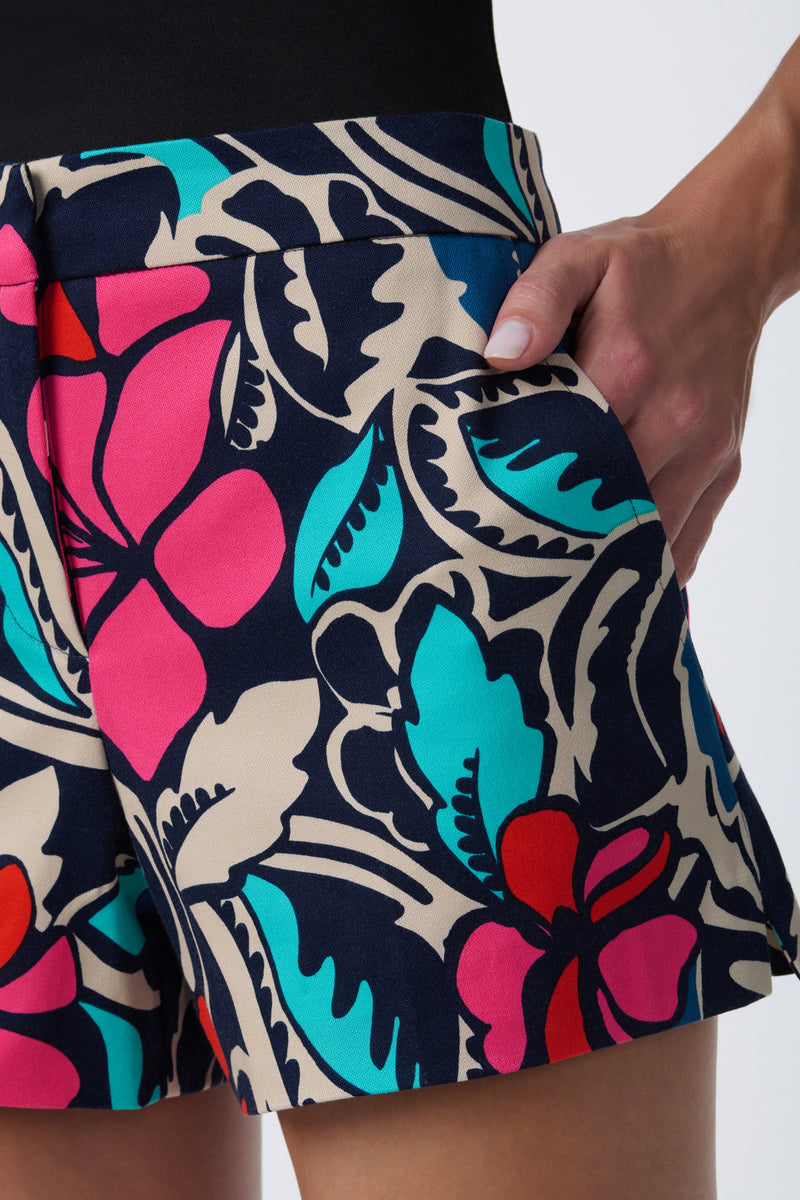 A person wearing the Corbin 2 Short, featuring a bold floral print in pink, teal, red, and navy, stands with one hand in their pocket against a plain light background.