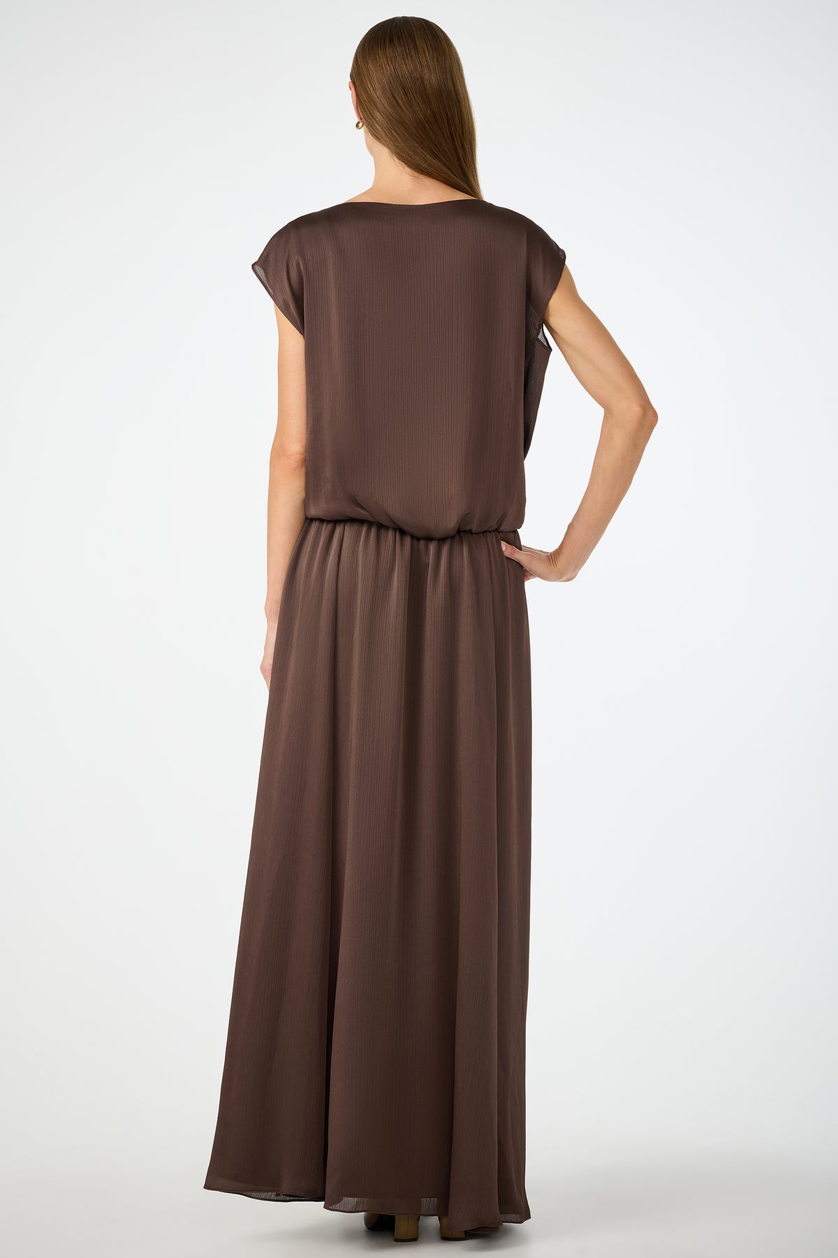 A woman with long, straight hair stands with her back to the camera, wearing the Vantage 2 Maxi Dress, a loose, short-sleeved, floor-length brown dress. The background is plain white.