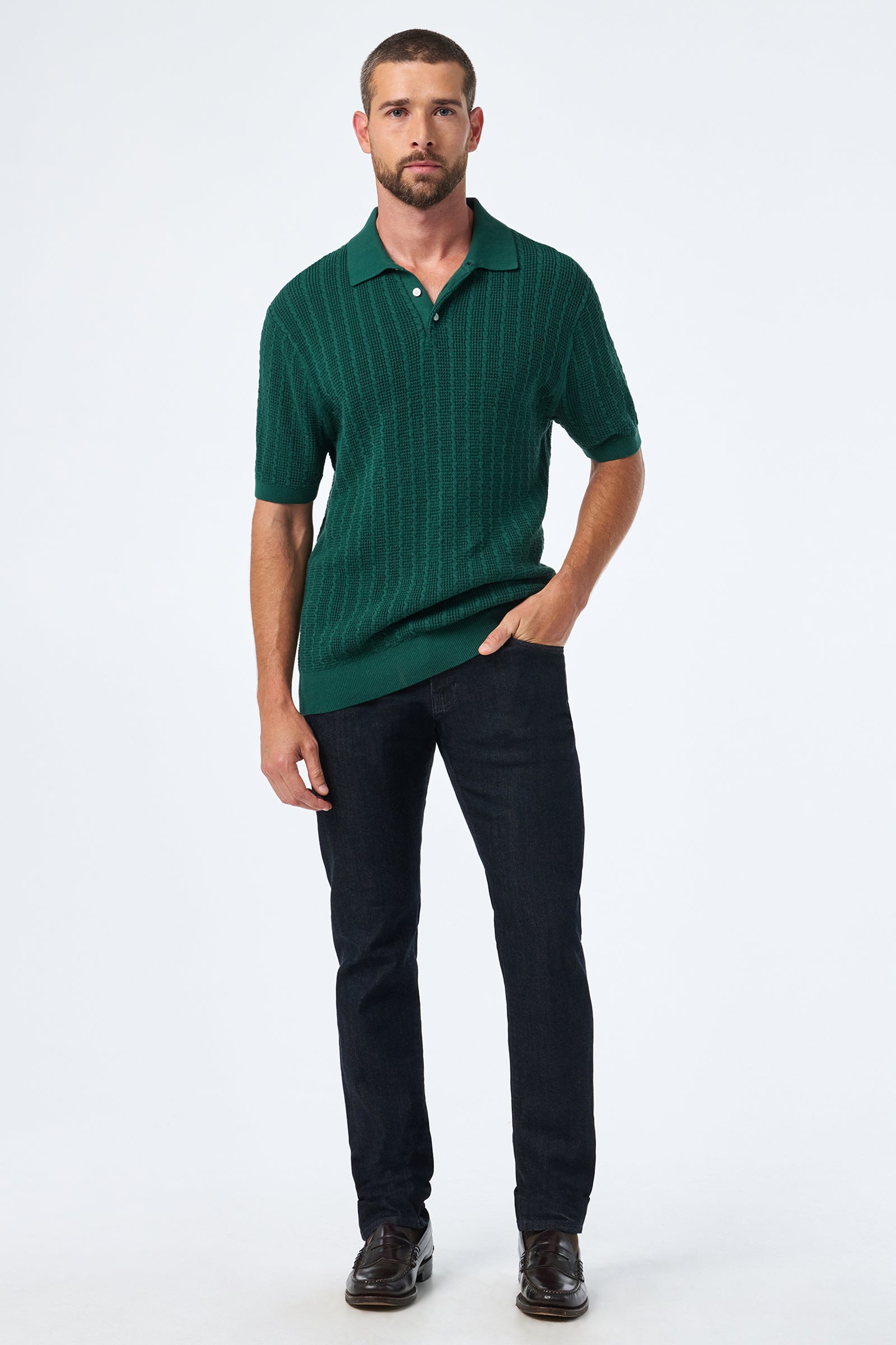 A man stands against a plain white background, wearing the Asher Ss Polo in green, dark jeans, and brown shoes, with one hand in his pocket and a neutral expression.