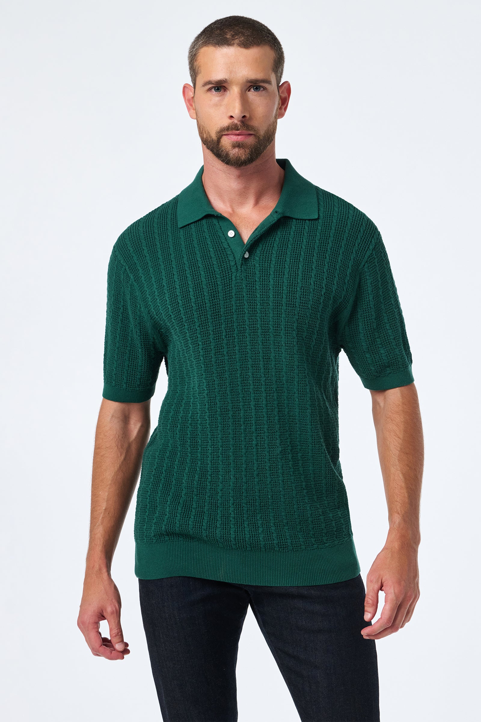 A man with short hair and a beard wears the Asher Ss Polo in textured dark green with dark jeans, standing against a plain light background.