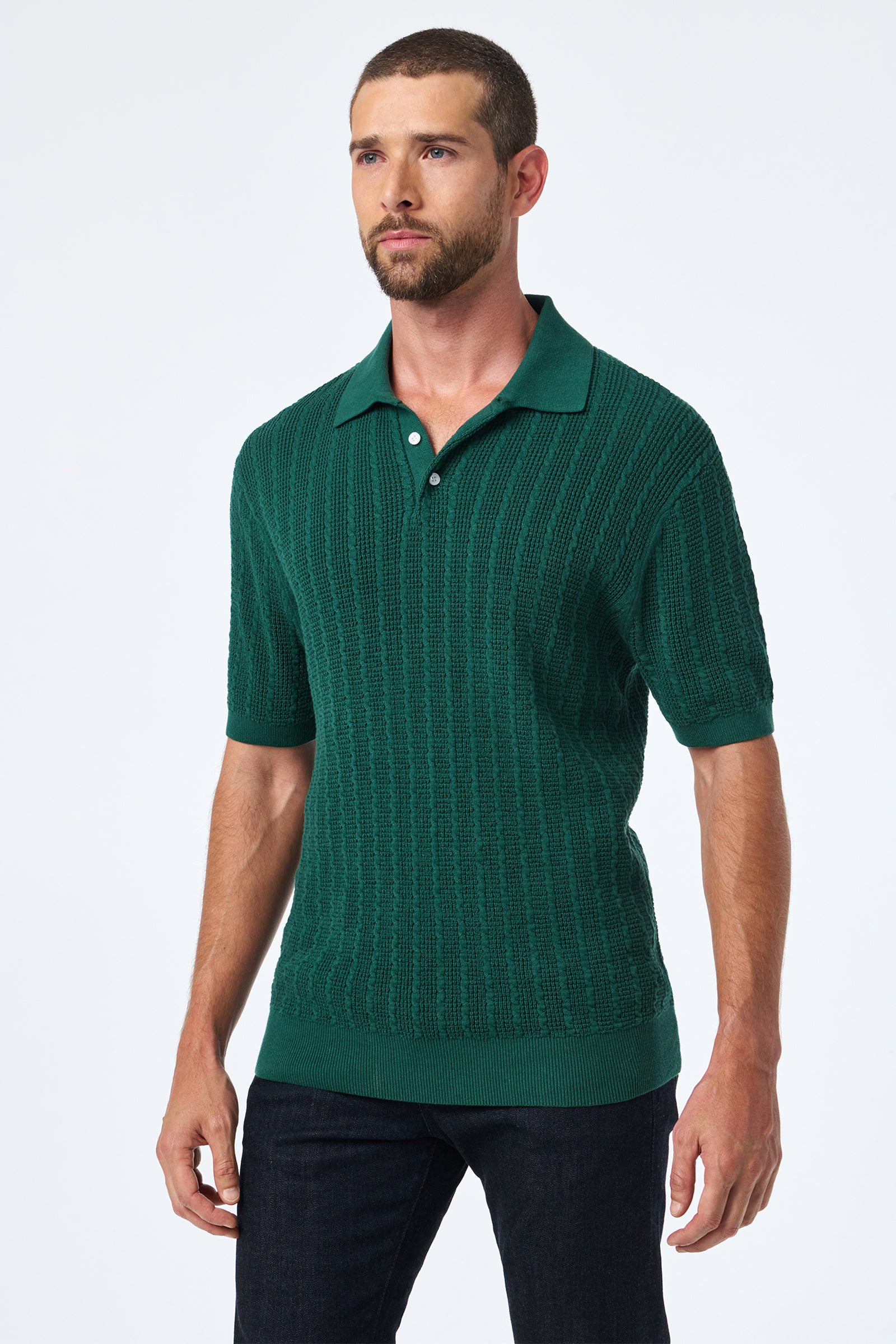 A man with short brown hair and a beard wears the Asher Ss Polo, a textured dark green short-sleeve knit shirt, with dark jeans, standing against a plain white background.