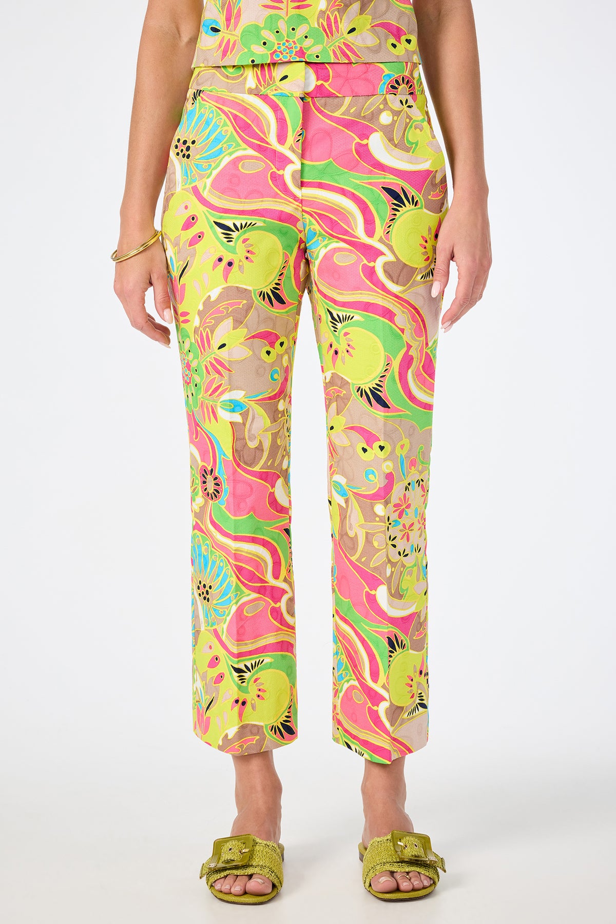 Person wearing Lulu Cropped Pant in the Venus color, featuring a psychedelic floral pattern with yellow, green, pink, and brown tones, paired with green slide sandals and a gold bracelet. Photo cropped at the shoulders.
