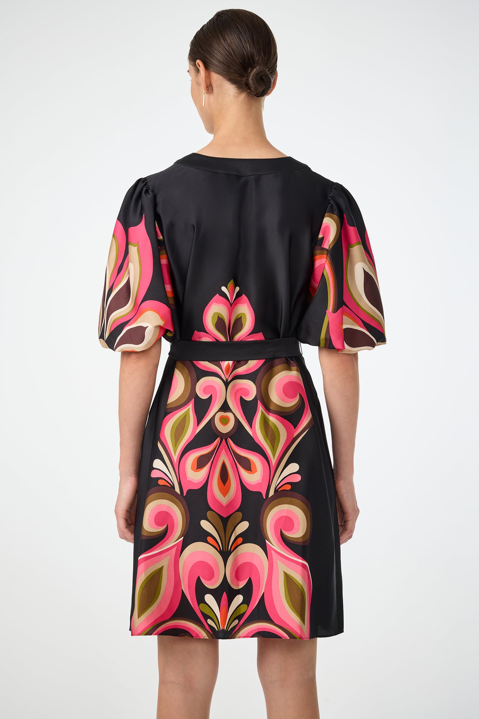 A woman with her hair in a bun is seen from the back wearing the Certonia Puff Sleeve Dress by Sanctuary—a short black dress featuring bold pink, orange, green, and brown patterns, puffed sleeves, and a belt.