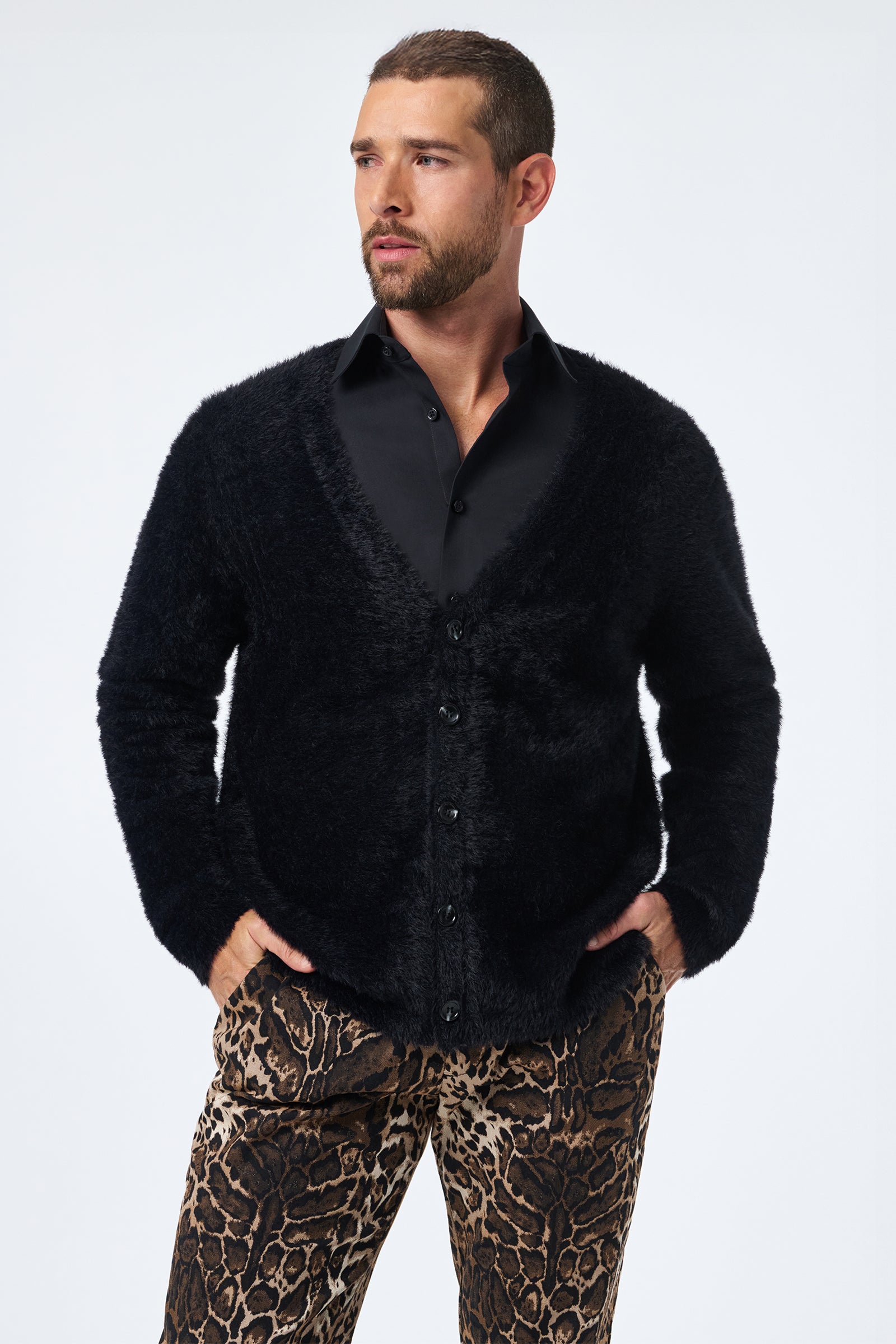 A man with short hair and a beard wears the Scott Fuzzy Cardigan in black over a black dress shirt and animal-print pants, standing with hands in pockets and gazing to the side against a plain light background.