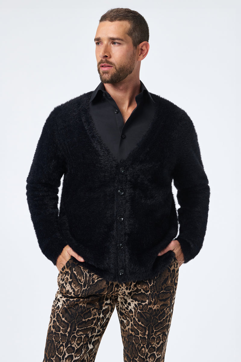A man with short hair and a beard wears the Scott Fuzzy Cardigan in black over a black dress shirt and animal-print pants, standing with hands in pockets and gazing to the side against a plain light background.