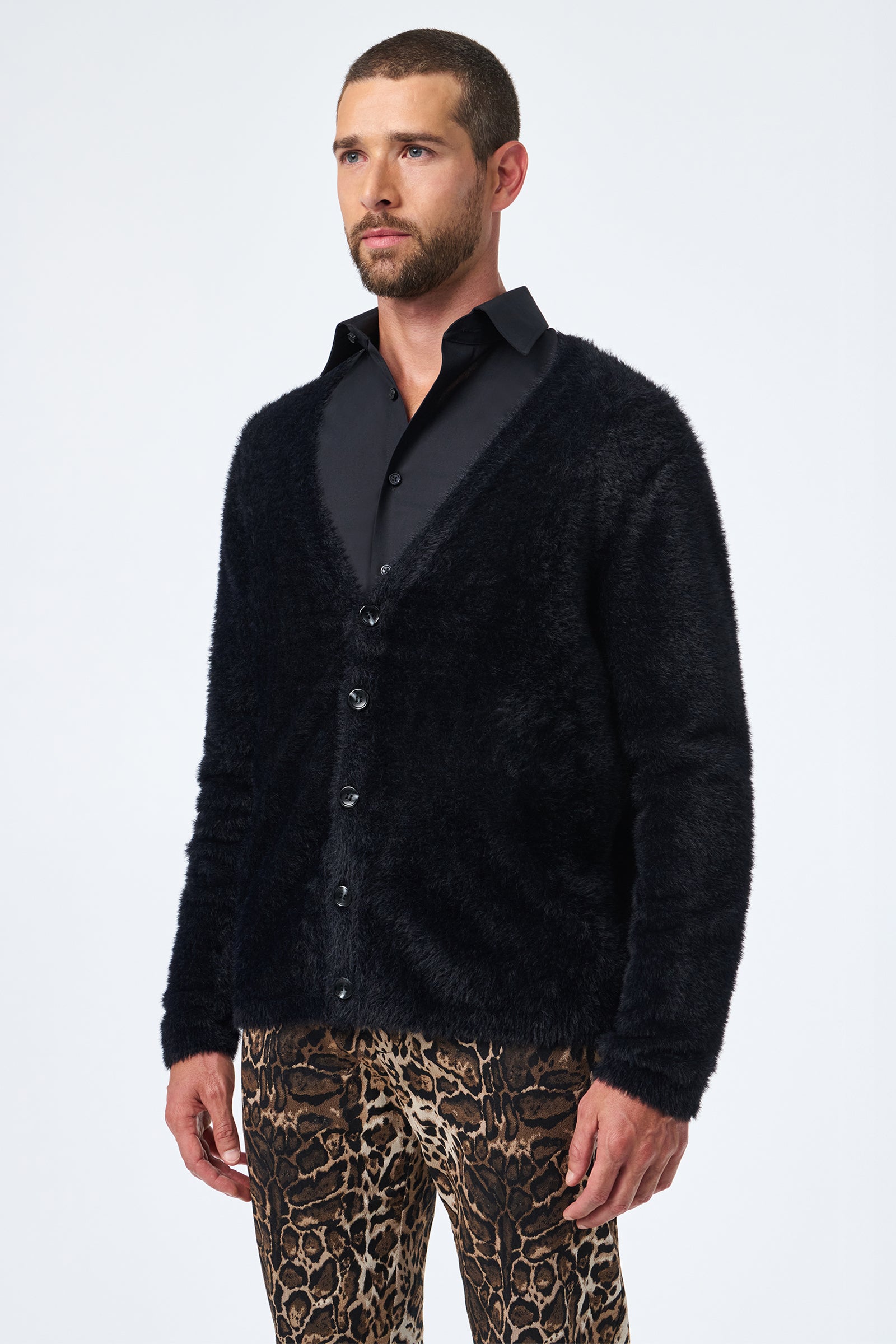 A man with short hair and a beard wears the Scott Fuzzy Cardigan in black over a black shirt and leopard print pants, standing against a plain white background.