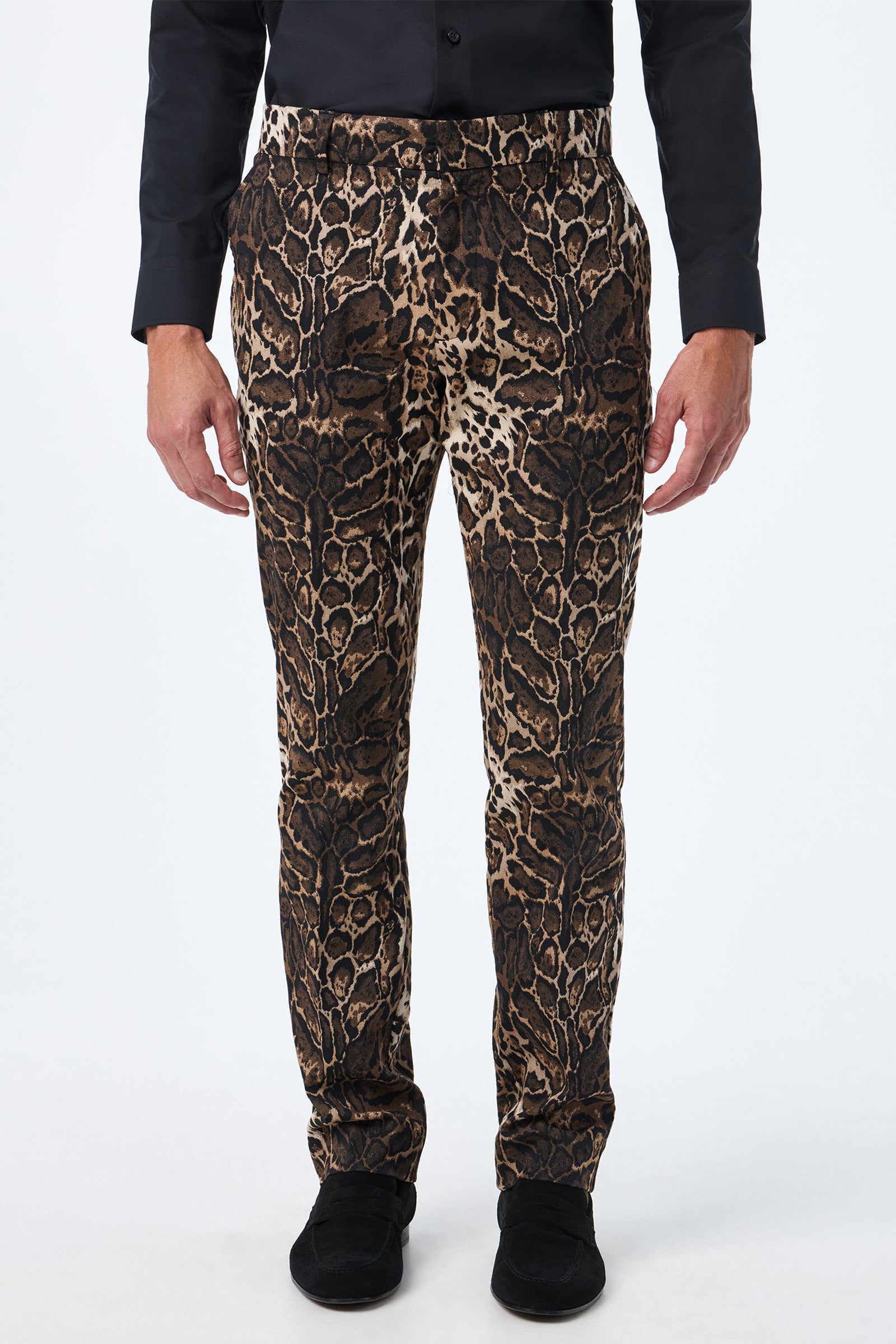 A person wearing the Clyde Slim Trouser - Lompoul Leopard, featuring a brown and black leopard print, paired with a black button-up shirt and black shoes, stands against a plain white background.