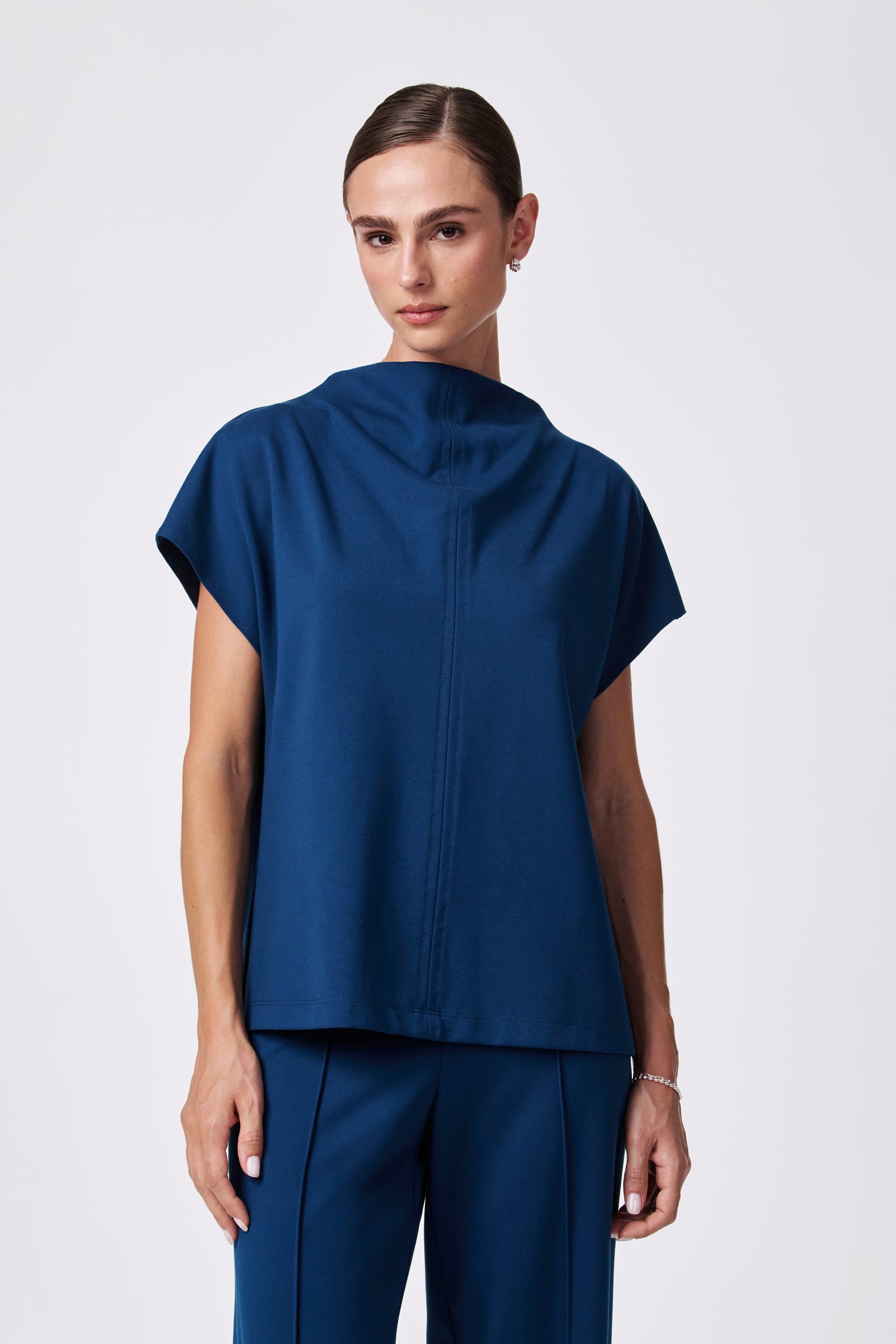 A woman with brown hair pulled back wears the loose-fitting, short-sleeved Pavilion Top in dark blue and matching pants, standing against a plain light background.