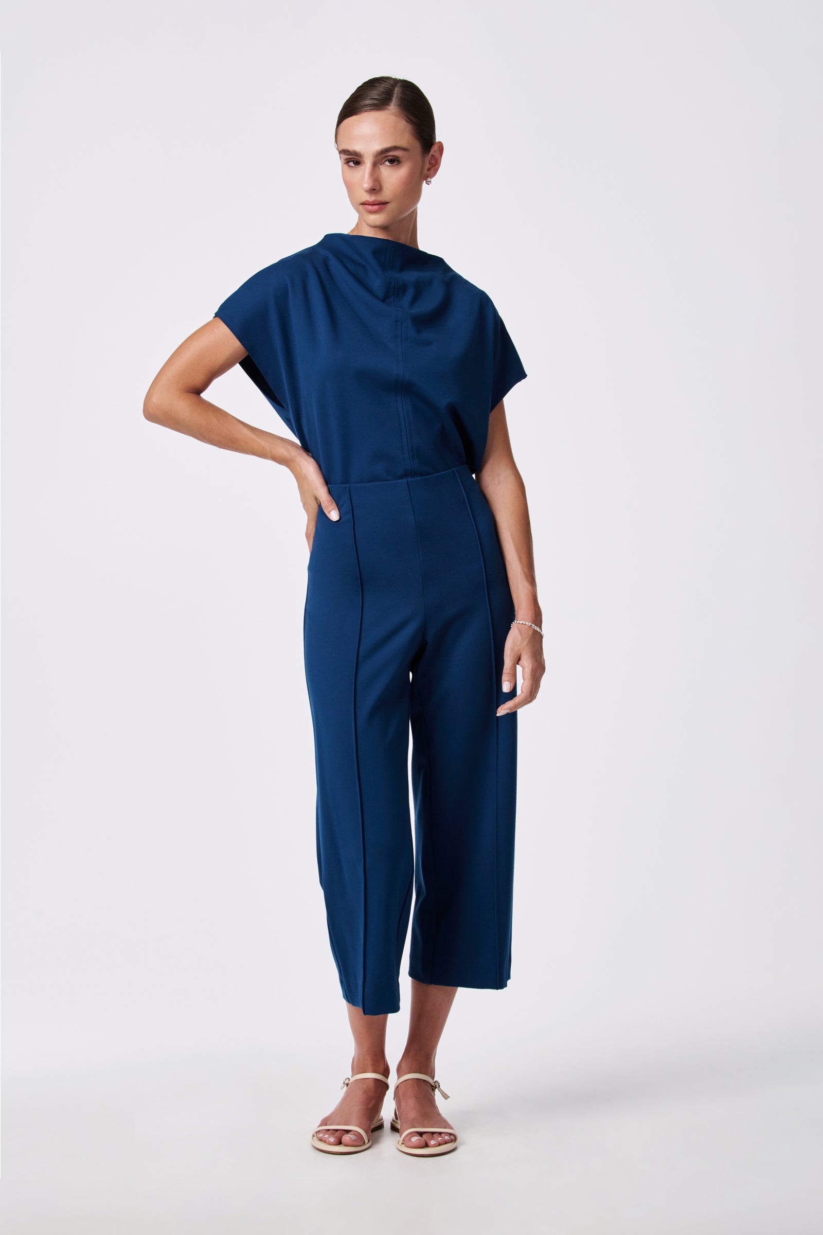 A woman wears the Santo Pant—high-waisted, cropped dark blue pants—with a matching short-sleeve draped-neckline top against a plain white background. She pairs the look with white sandals and has her hair pulled back.