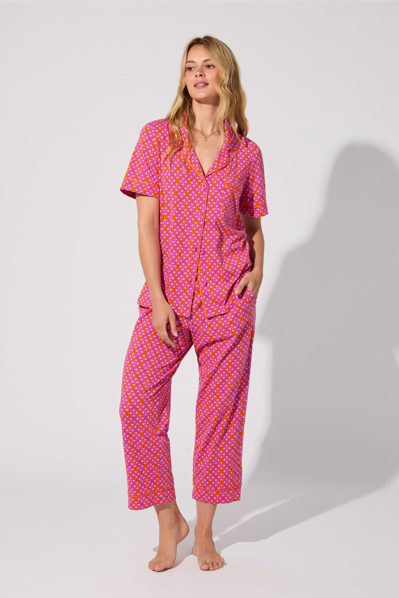 A woman stands barefoot against a plain background, wearing the Crop Classic PJ Set in Chain Geo. The set features a short-sleeve button-up shirt and loose-fitting pants with a geometric chain print.
