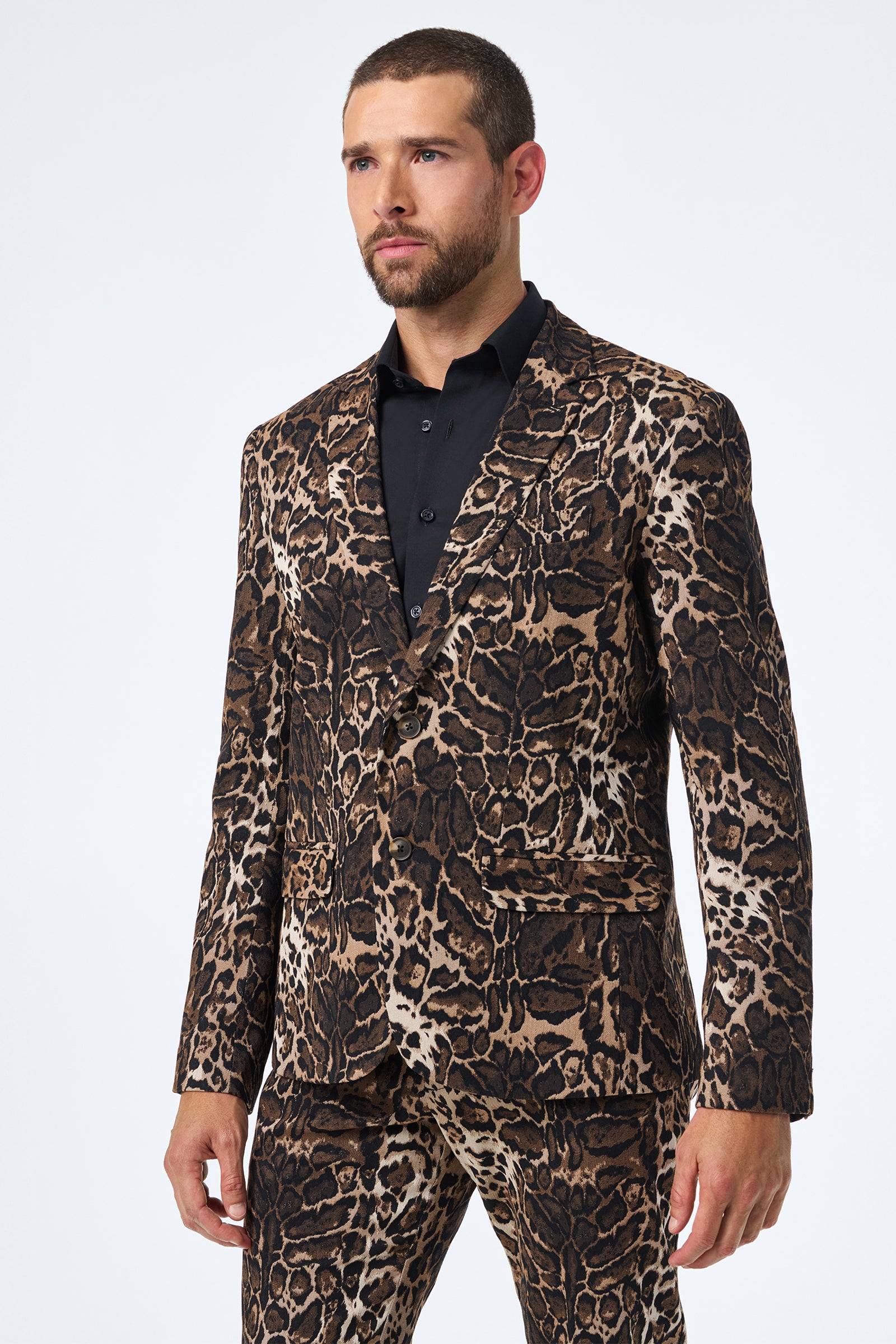 A man with short hair and a beard models the Thurston Blazer - Lompoul Leopard, a bold animal-print suit in brown, black, and tan over a black dress shirt, standing against a plain white background.