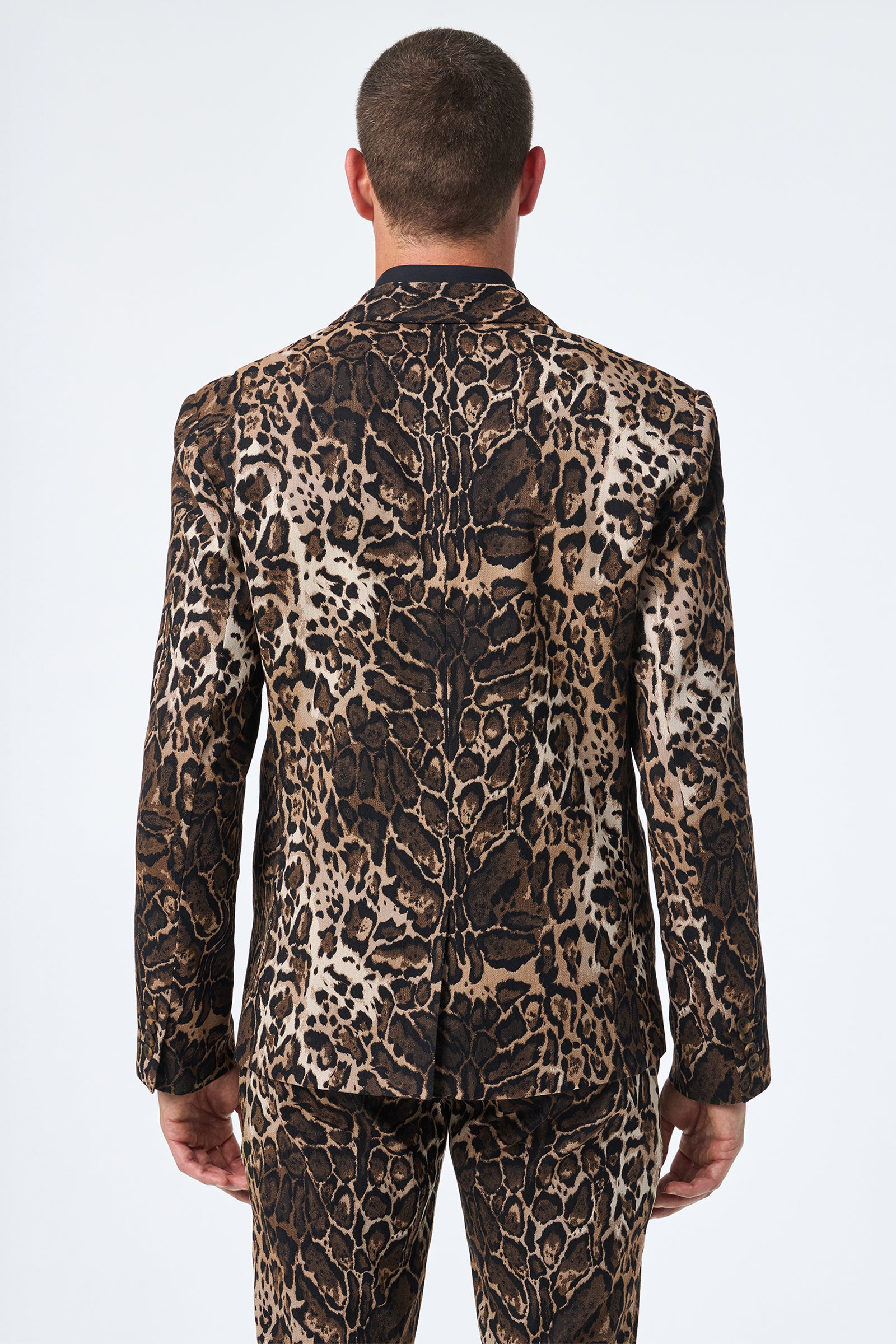 A person with short hair is seen from behind wearing the Thurston Blazer - Lompoul Leopard, featuring a bold brown, black, and tan animal print pattern, paired with matching pants against a plain white background.