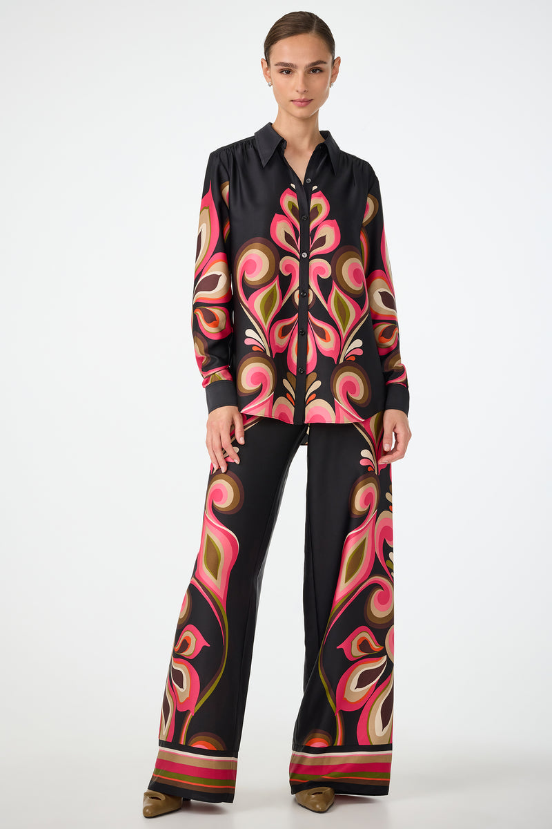 A woman wearing the Cassavetes Border Shirt - Sanctuary poses against a white background, paired with wide-leg pants featuring bold paisley prints in pink, orange, and beige. She has her hair tied back and wears brown shoes.