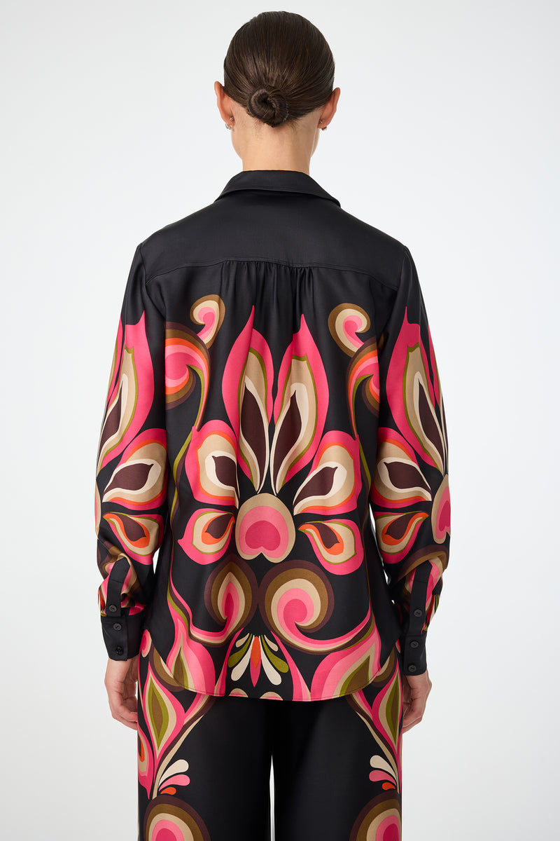 A person with dark hair in a low bun is shown from the back wearing the Cassavetes Border Shirt - Sanctuary, a black shirt and pants set with a bold, retro-inspired pink, brown, cream, and olive pattern against a plain background.