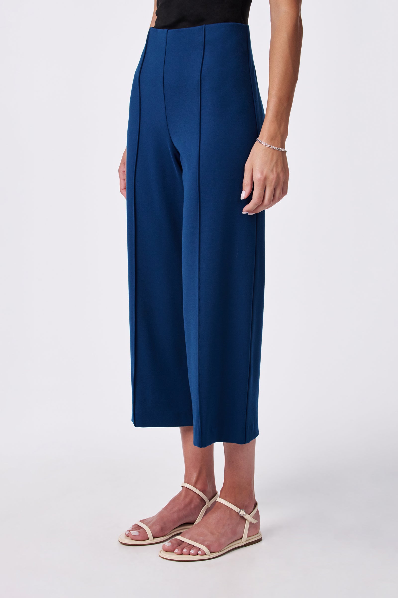 A person stands against a plain white background wearing the Santo Pant, a pair of high-waisted, wide-leg blue cropped pants, styled with a sleeveless black top, white strappy flat sandals, and a delicate bracelet.