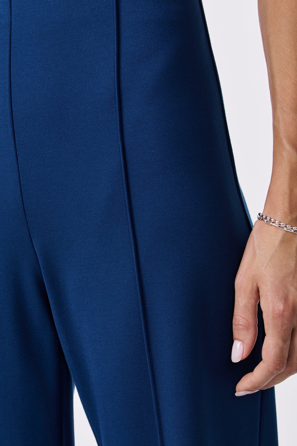 Close-up of a person wearing the Santo Pant in blue, highlighting the side seam. Part of their hand with a silver chain bracelet and manicured nails is visible against a plain, light background.