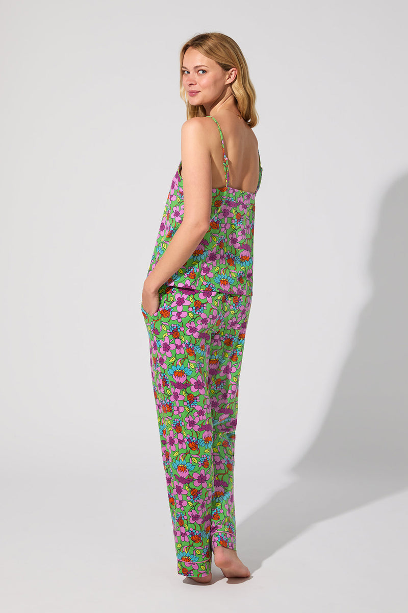 A woman stands barefoot, facing slightly away, wearing the Cami PJ Set - Garden Floral, which features a loose floral camisole top and matching pants. The background is plain and light-colored.