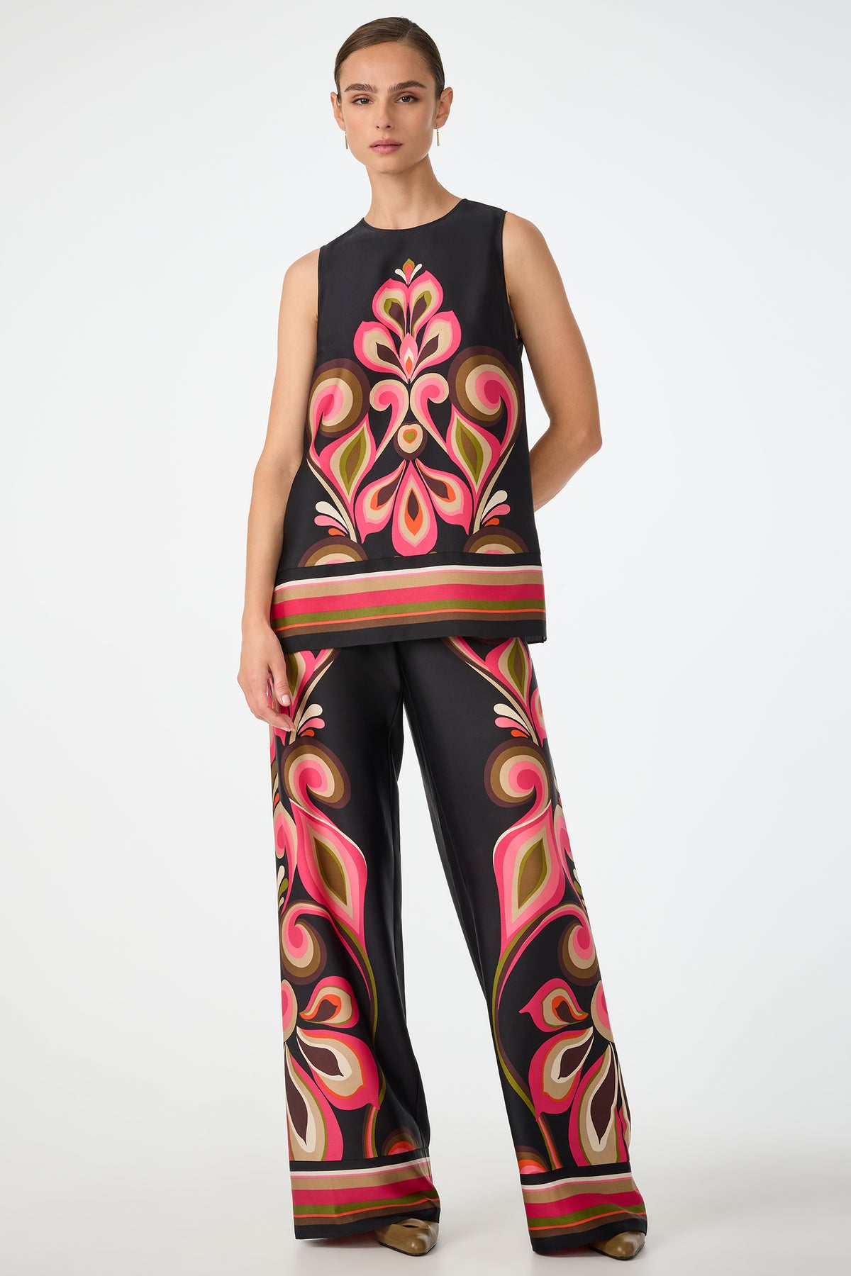 A woman poses against a plain backdrop wearing Verity Border Pants—wide-leg and sleeveless—with bold, colorful, symmetrical floral patterns in pink, red, green, and beige.