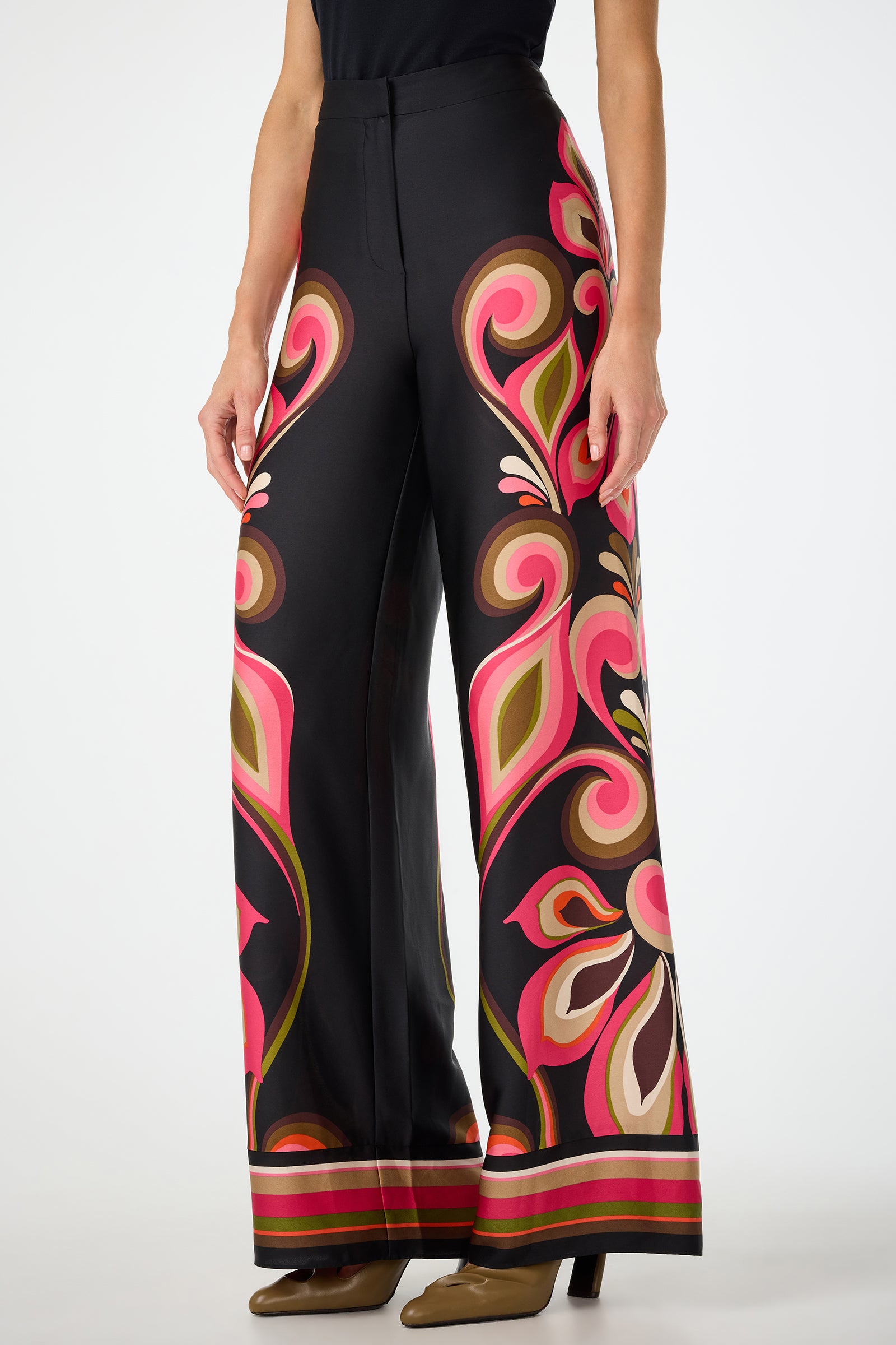 A woman wears the Verity Border Pants—high-waisted, wide-leg black pants with bold pink, green, red, and tan floral and swirl patterns—paired with a black top and brown heeled shoes.