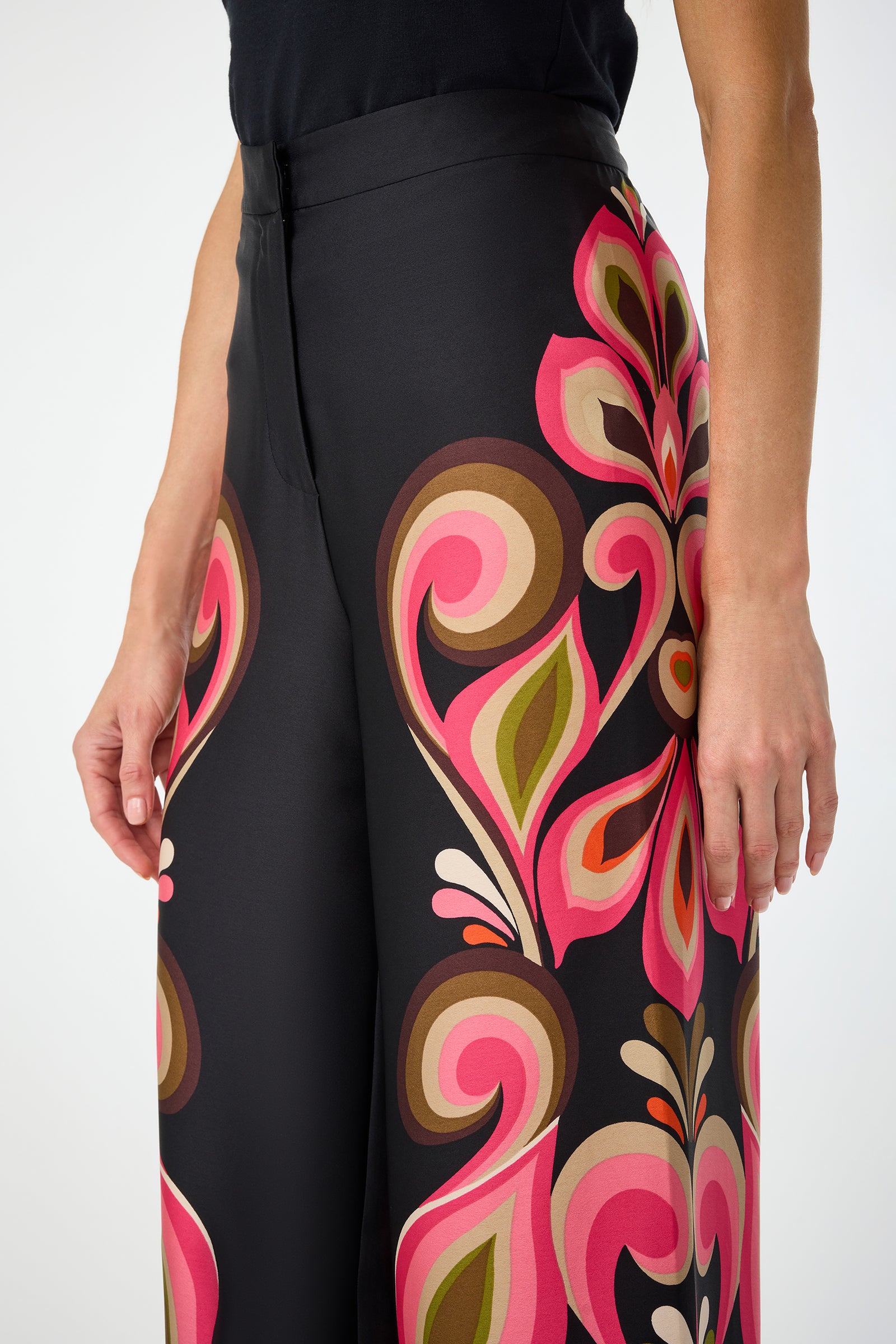 A person wears Verity Border Pants—high-waisted black trousers with a bold swirl pattern in pink, red, brown, and green—paired with a sleeveless black top. Only their torso and upper legs are visible.