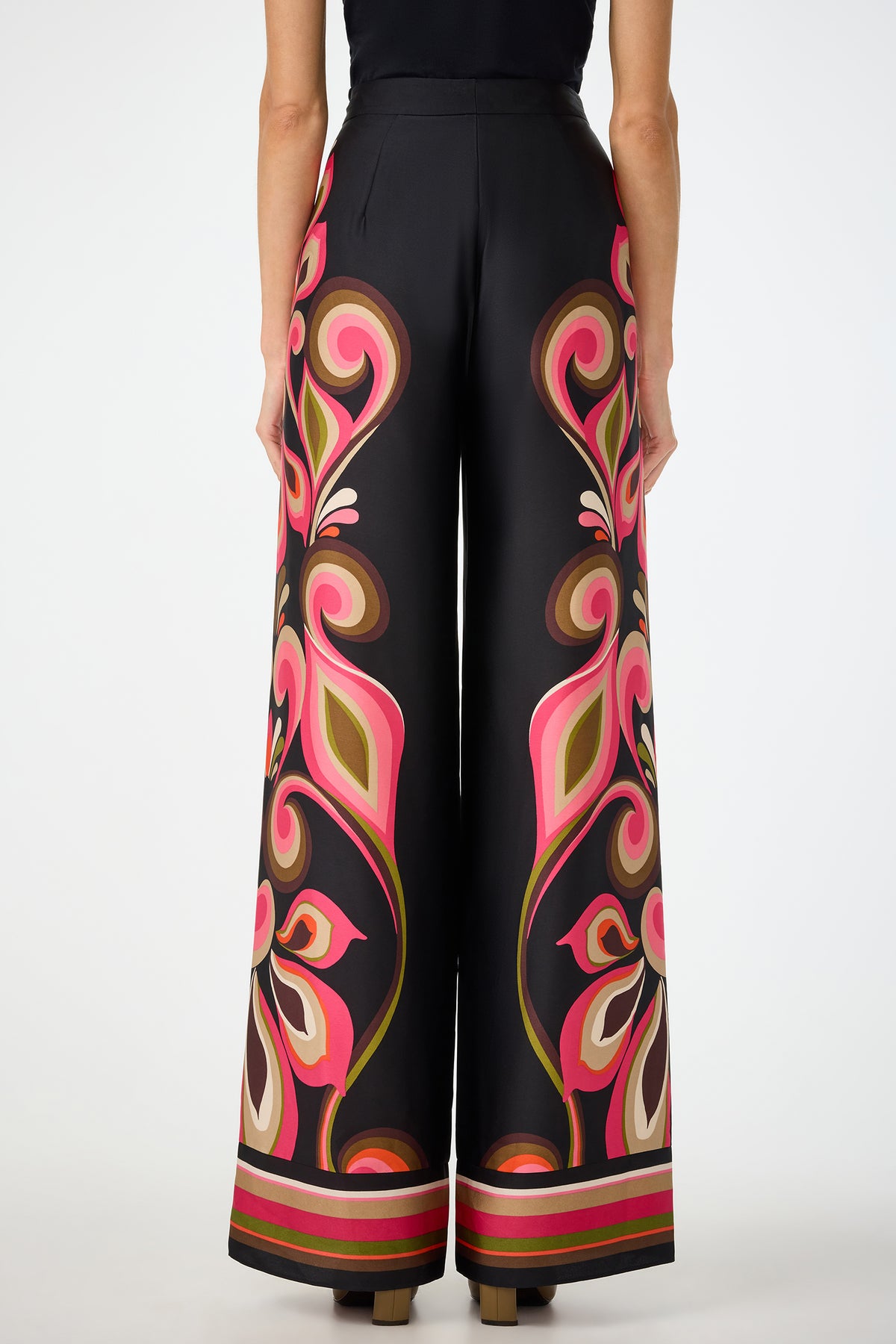 A woman is seen from the back against a plain background wearing Verity Border Pants—high-waisted, wide-leg black pants featuring bold floral and swirl patterns in pink, brown, olive, and beige.
